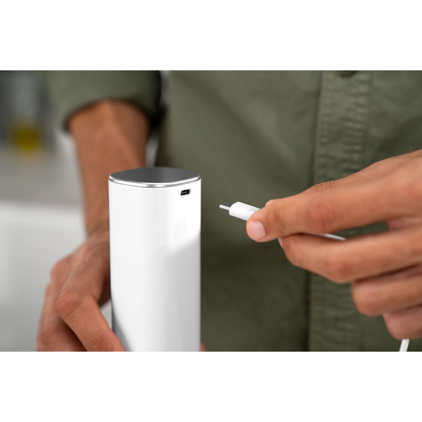 Ręka trzymająca biały kabel ładujący, gotowa podłączyć go do białego cylindrycznego urządzenia, na białej powierzchni.