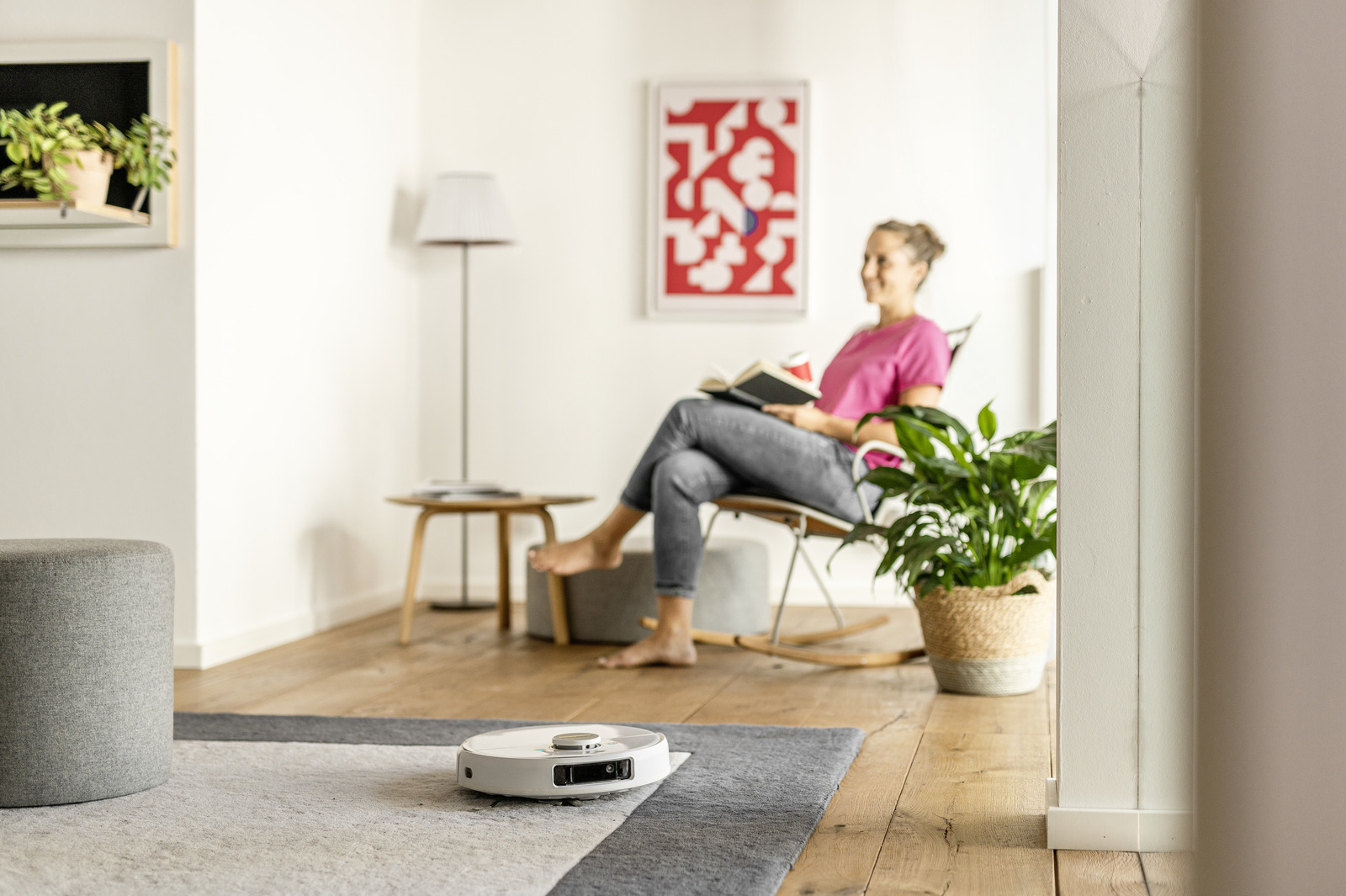 Kobieta czyta na krześle, robot sprząta podłogę, rośliny w pomieszczeniu w domowym otoczeniu.
