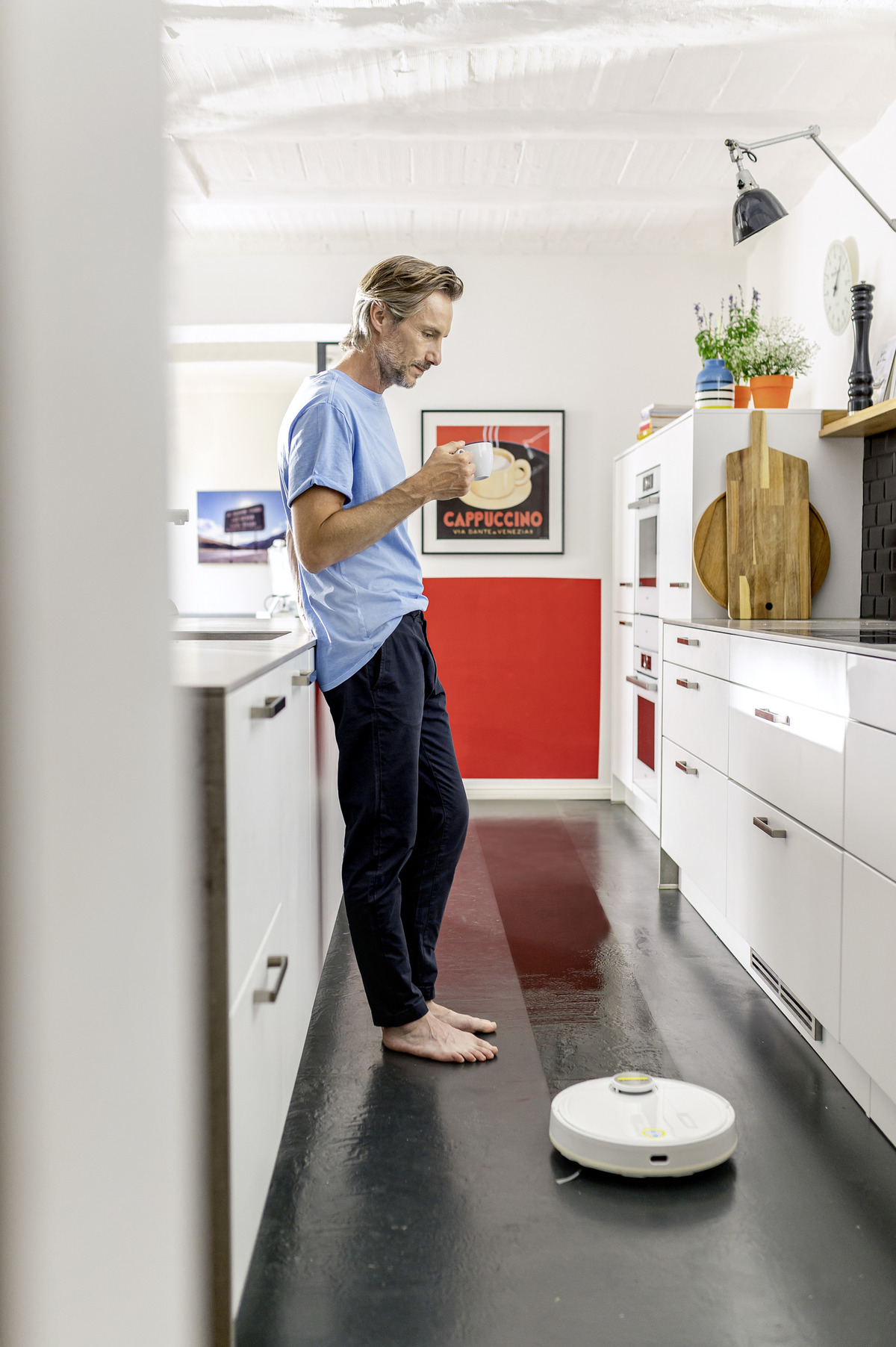 Un hombre en una cocina con suelo blanco, apoyado en una encimera, mirando una taza y una aspiradora robótica.