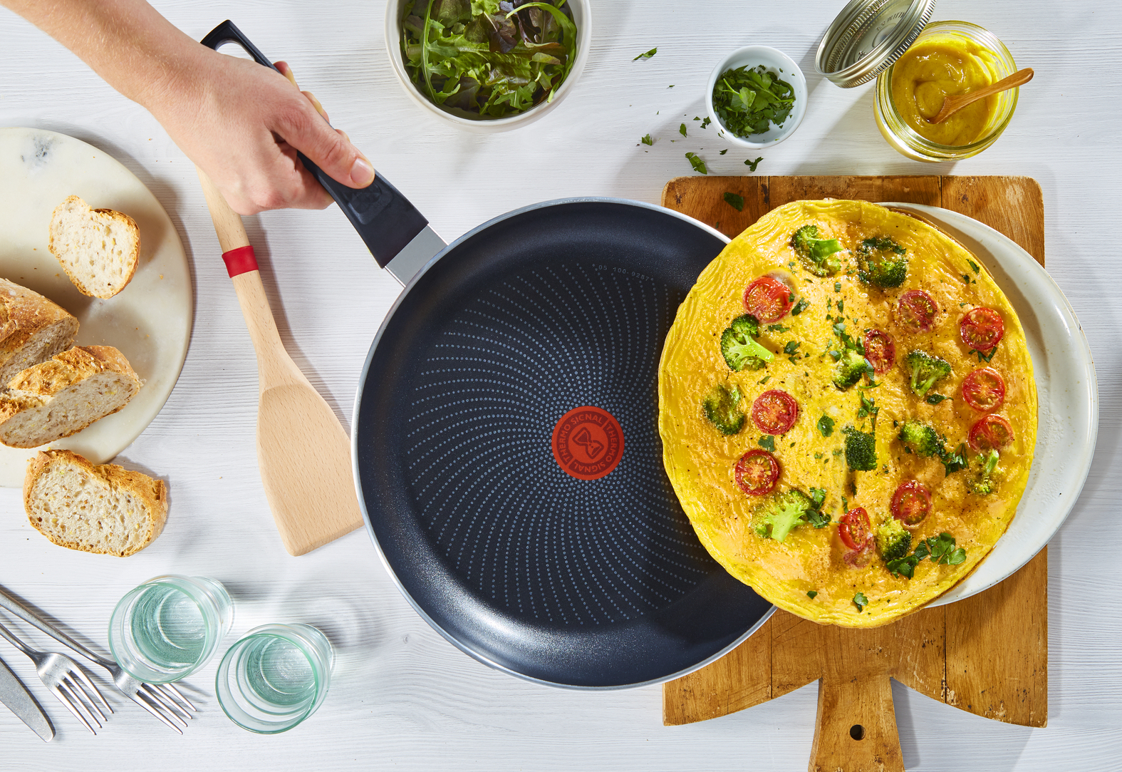 Overhead shot of a pan with omelet, wooden spatula, ingredients, and glasses on a table.
