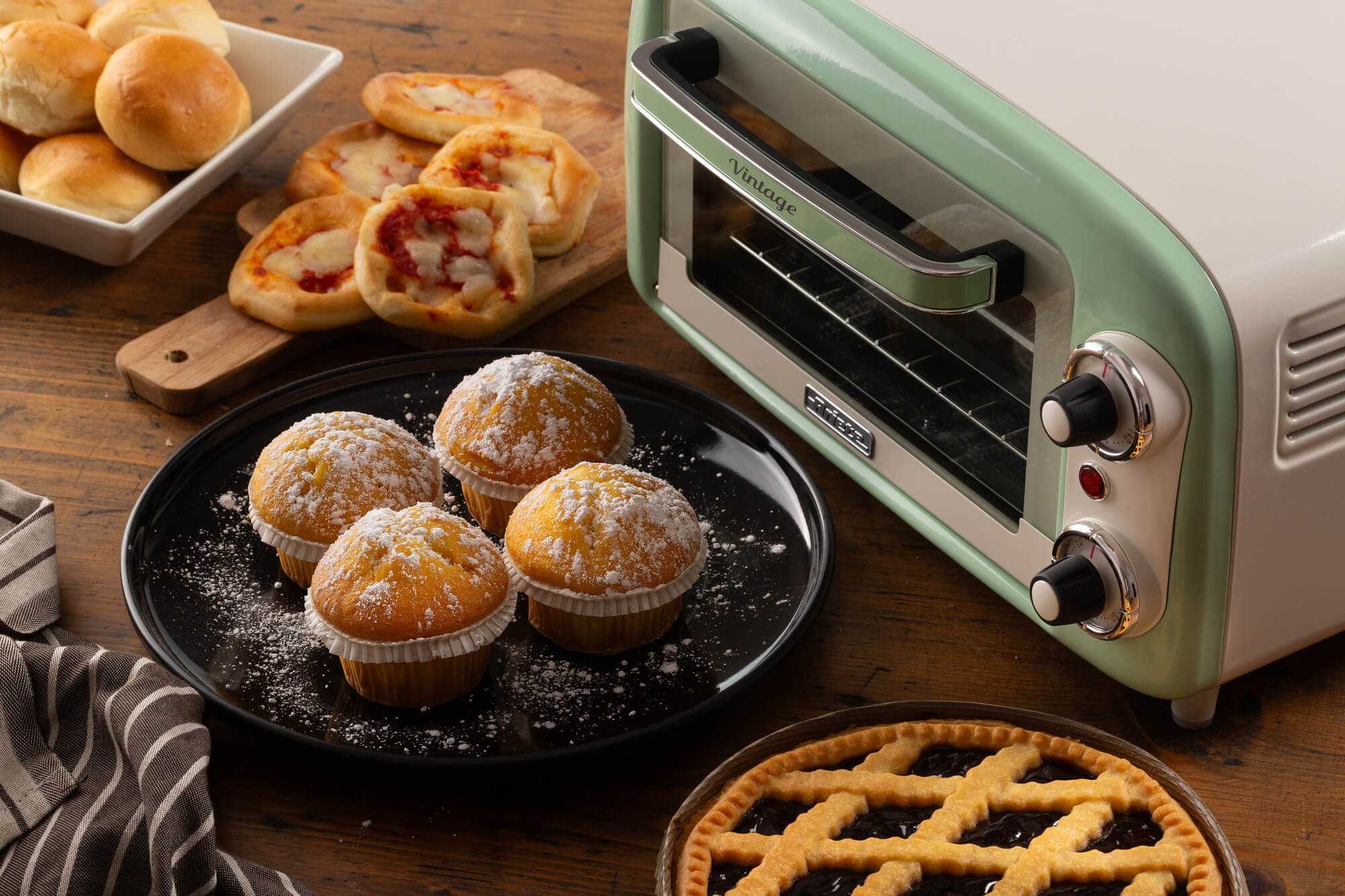 Zabytkowy zielono-biały piekarnik z wypiekami. Muffiny, pizze i ciasto na drewnianym stole.