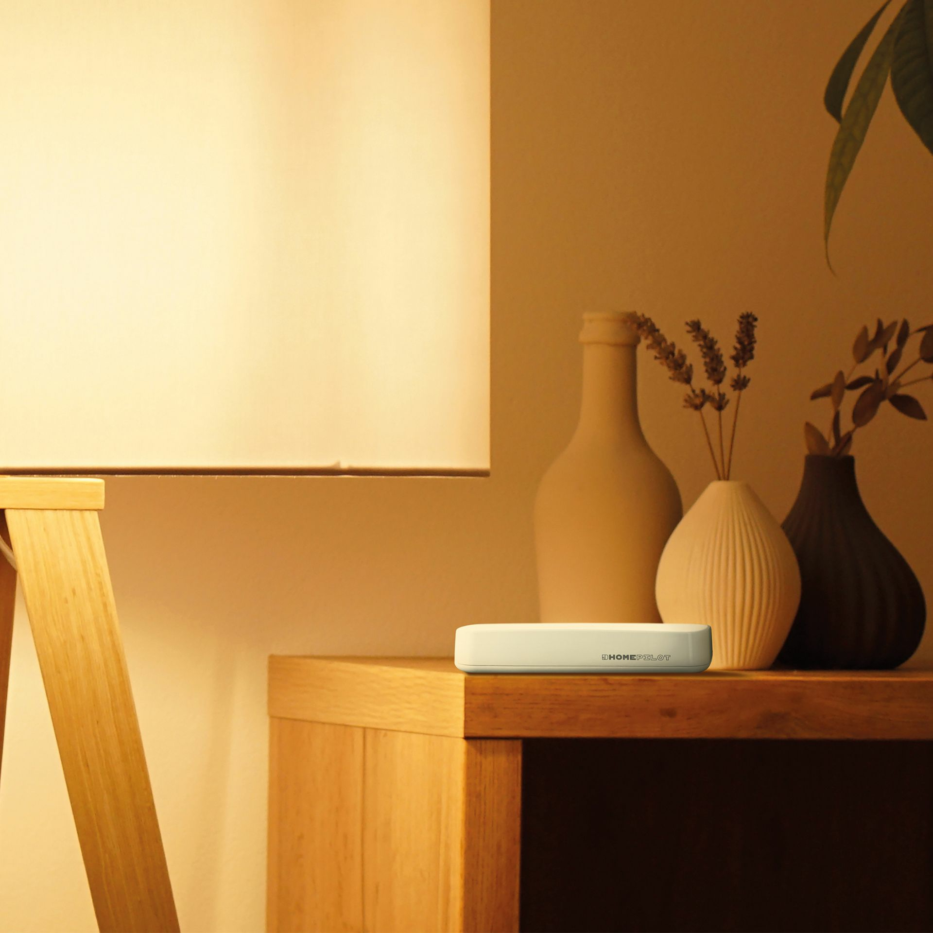 Ein kleines weißes Gerät liegt auf einer Holzoberfläche in der Nähe einer Lampe und Vasen, mit HOMEPILOT beschriftet.