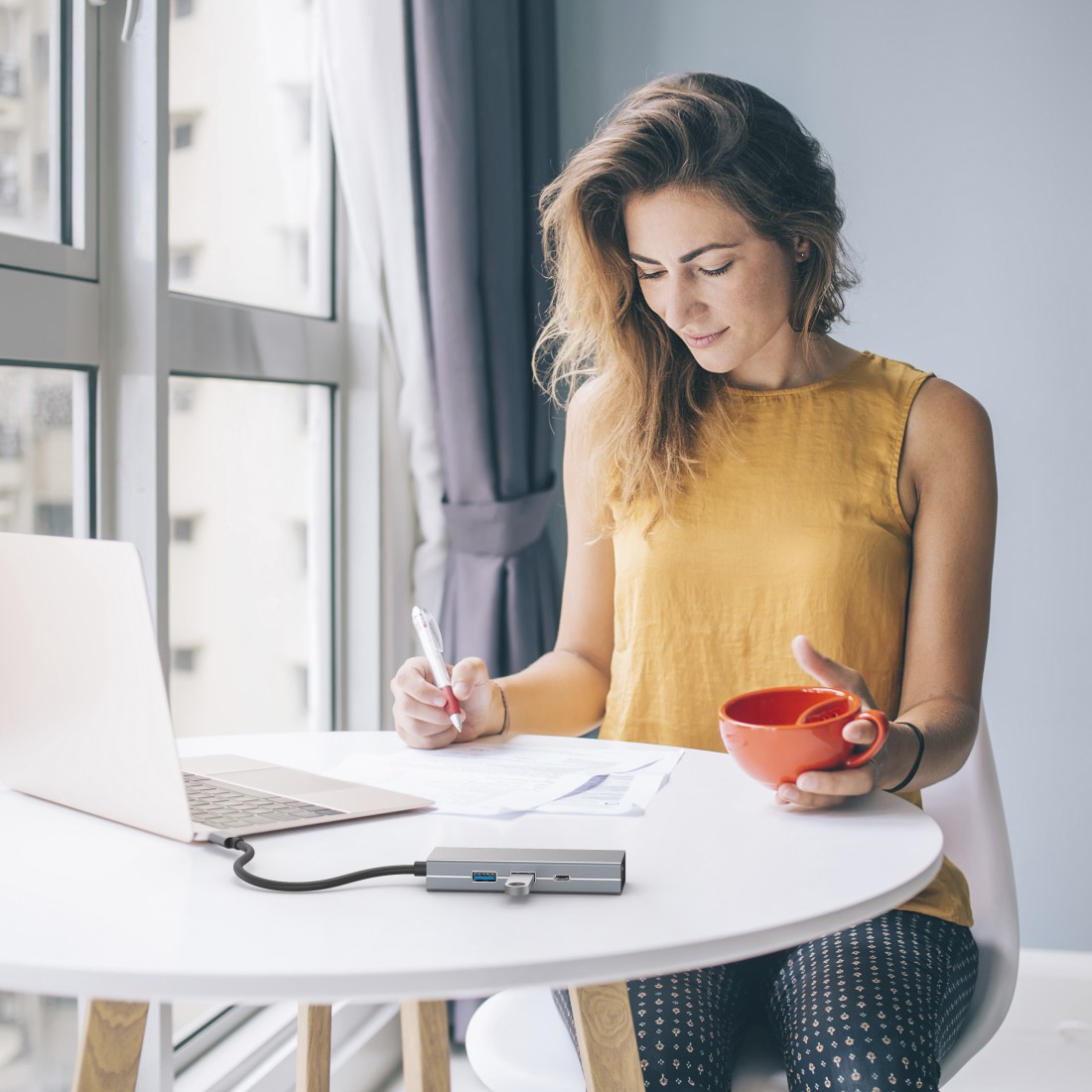 Kobieta pracuje przy stole z laptopem, papierami i czerwoną filiżanką.