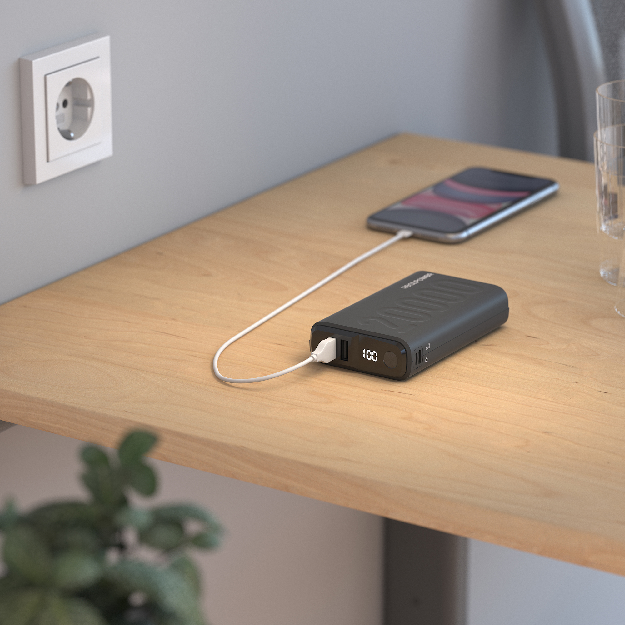 Eine Powerbank lädt ein Telefon auf einem Holztisch in der Nähe einer Steckdose und einer Pflanze.