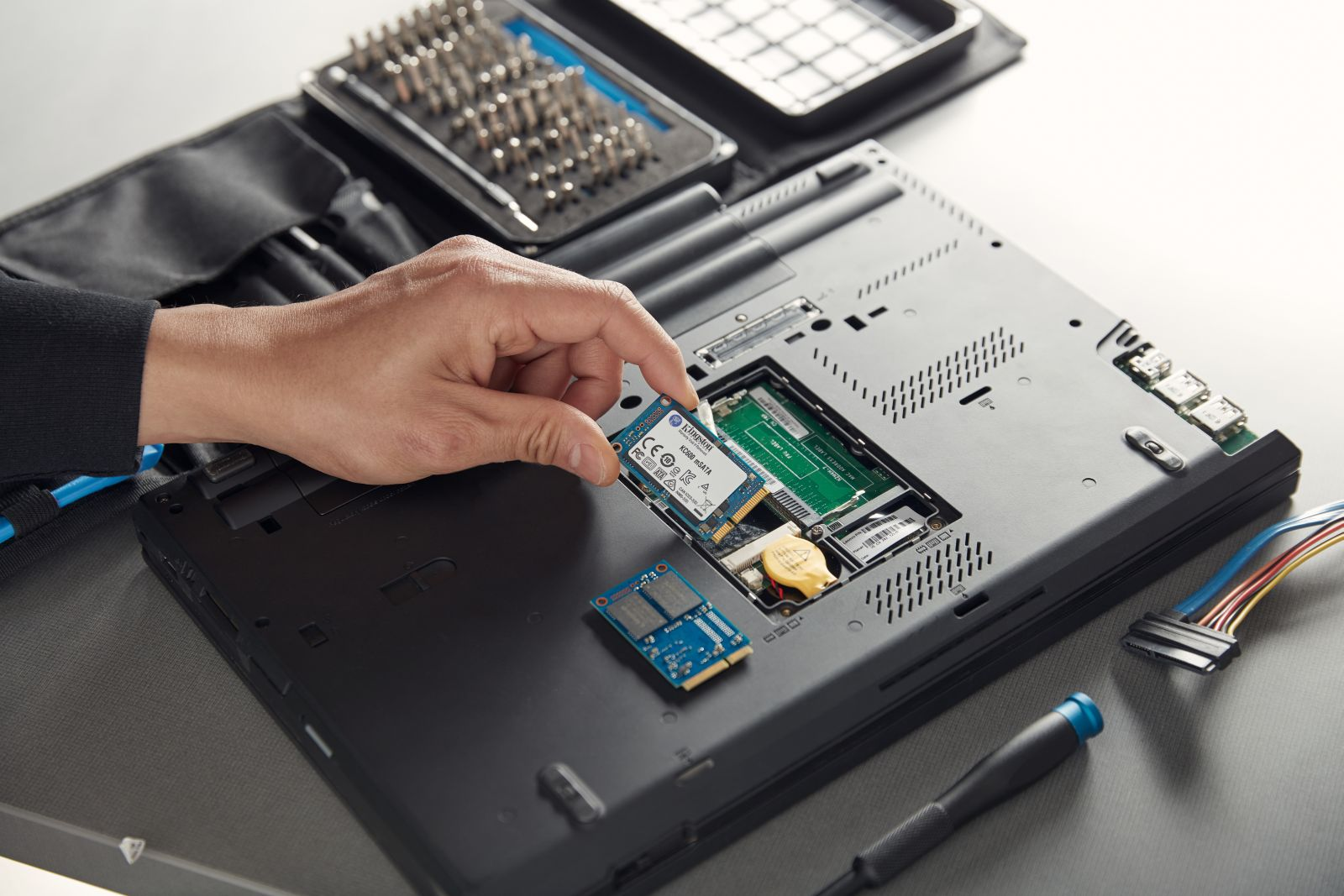 Ręka osoby instalującej dysk SSD w laptopie. Widoczne śrubokręty i inne komponenty.