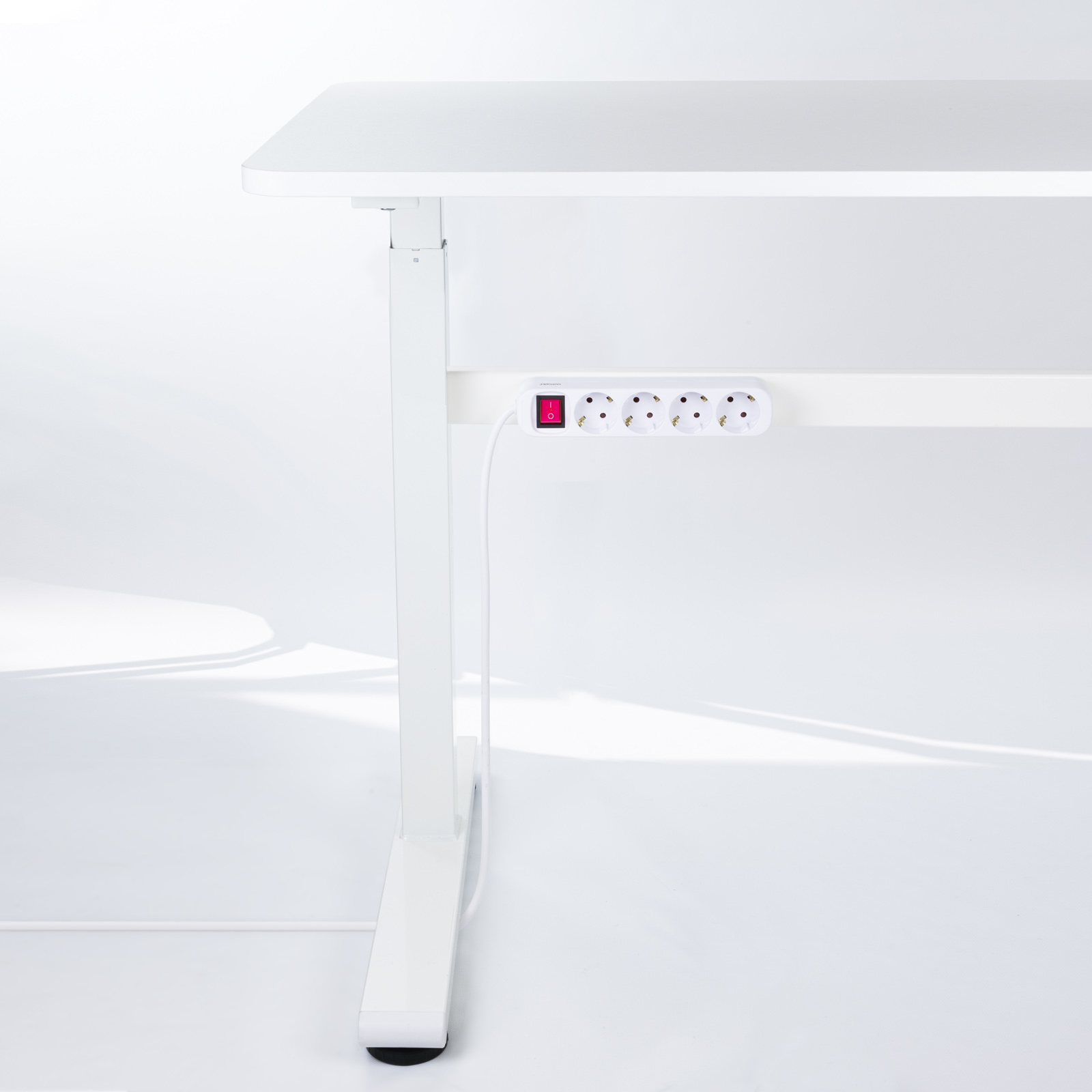 Białe biurko z podłączoną listwą zasilającą. Widoczny czerwony przełącznik. Biała ściana w tle.