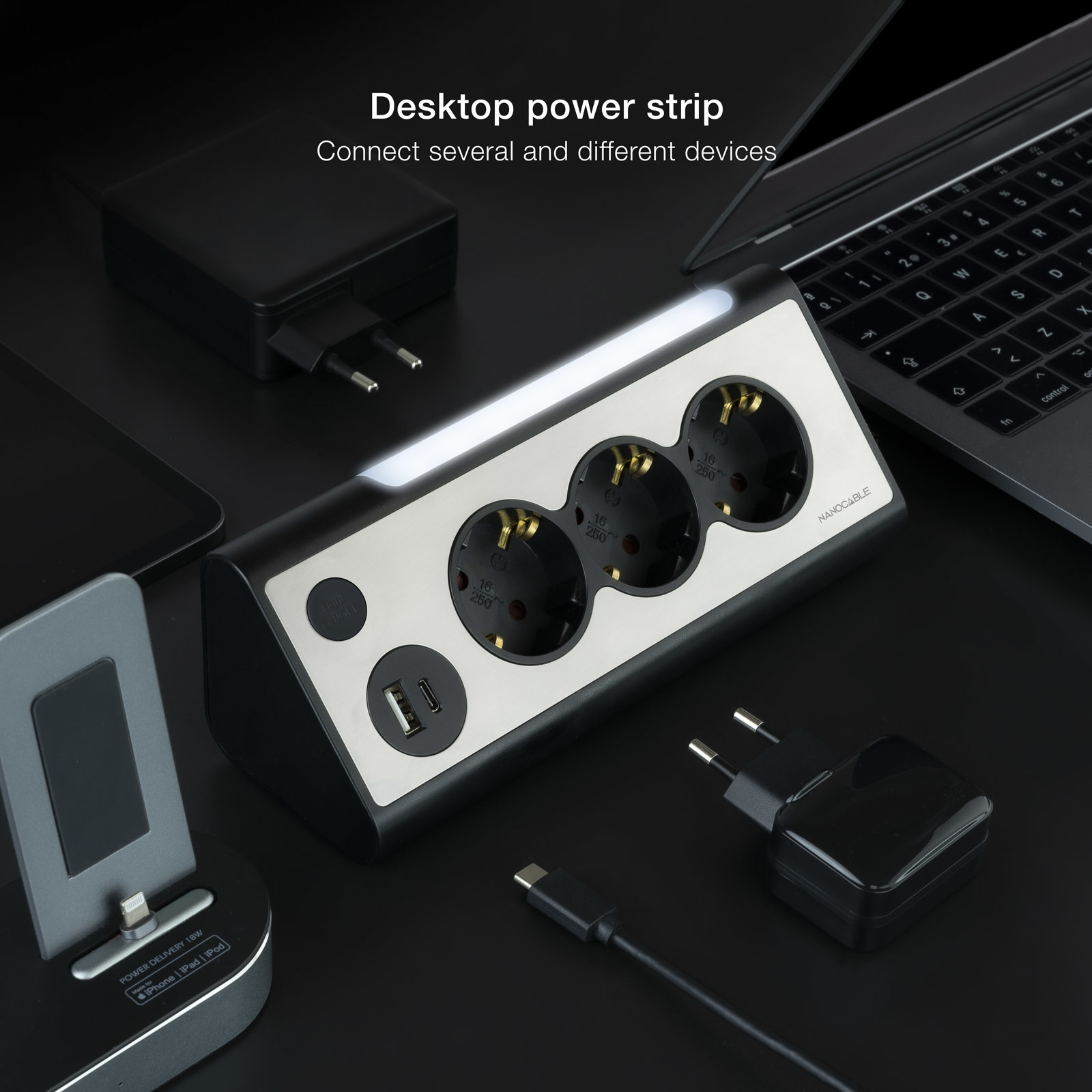 Een zwart-zilveren stekkerdoos met stopcontacten, USB-poorten en een lampje. Een laptop en opladers staan in de buurt.