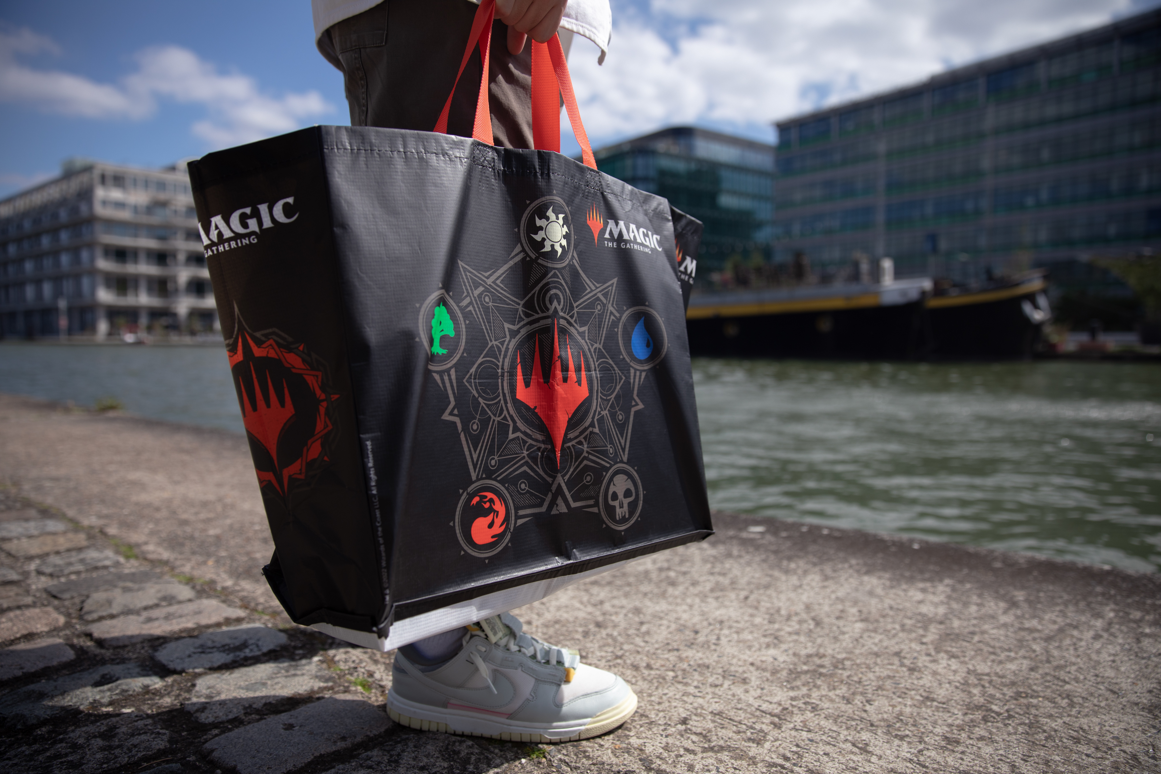 Persona sosteniendo una bolsa negra de Magic: The Gathering cerca del agua. Edificios al fondo.