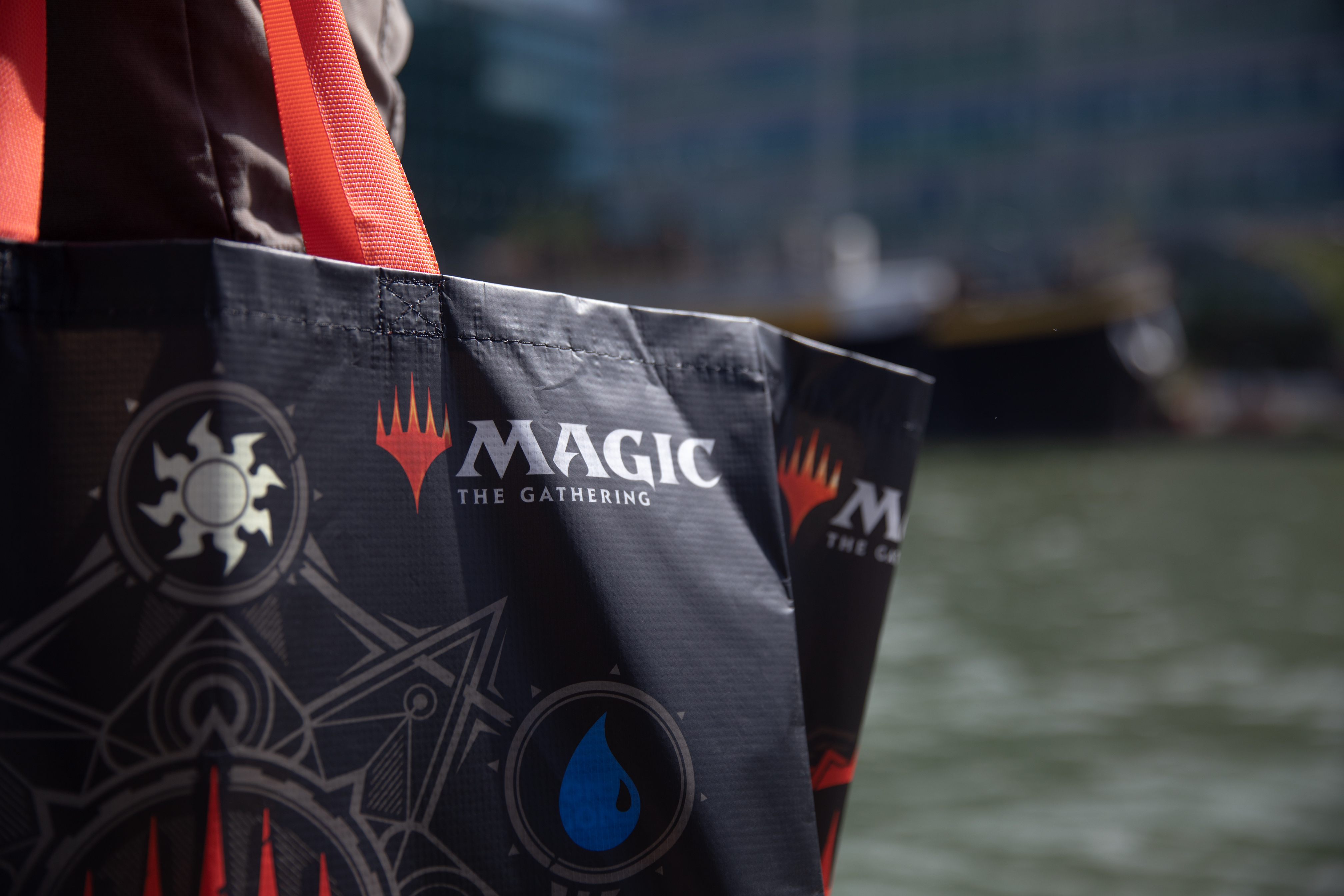 Persona sosteniendo una bolsa 'Magic: The Gathering' negra, agua y barco en el fondo.