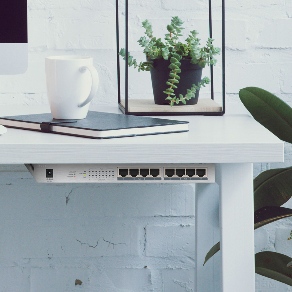 Przełącznik sieciowy zainstalowany pod białym biurkiem z rośliną w tle.