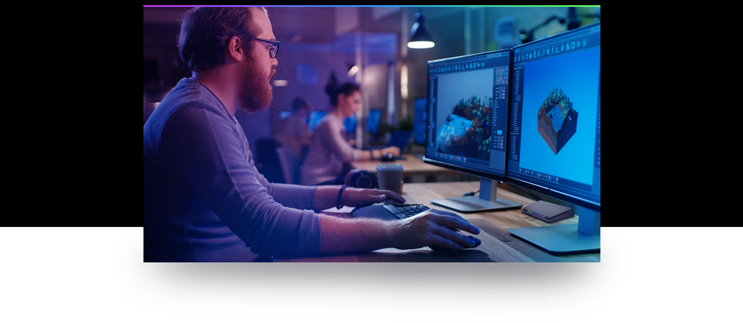 Mężczyzna w okularach używa komputera z dwoma monitorami. Kobieta pracuje w tle.