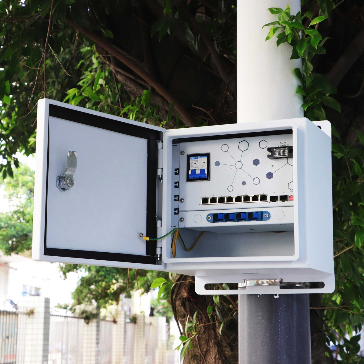 Biała zewnętrzna skrzynka sieciowa zamontowana na słupie, otwarta, pokazująca komponenty i okablowanie, z drzewem.