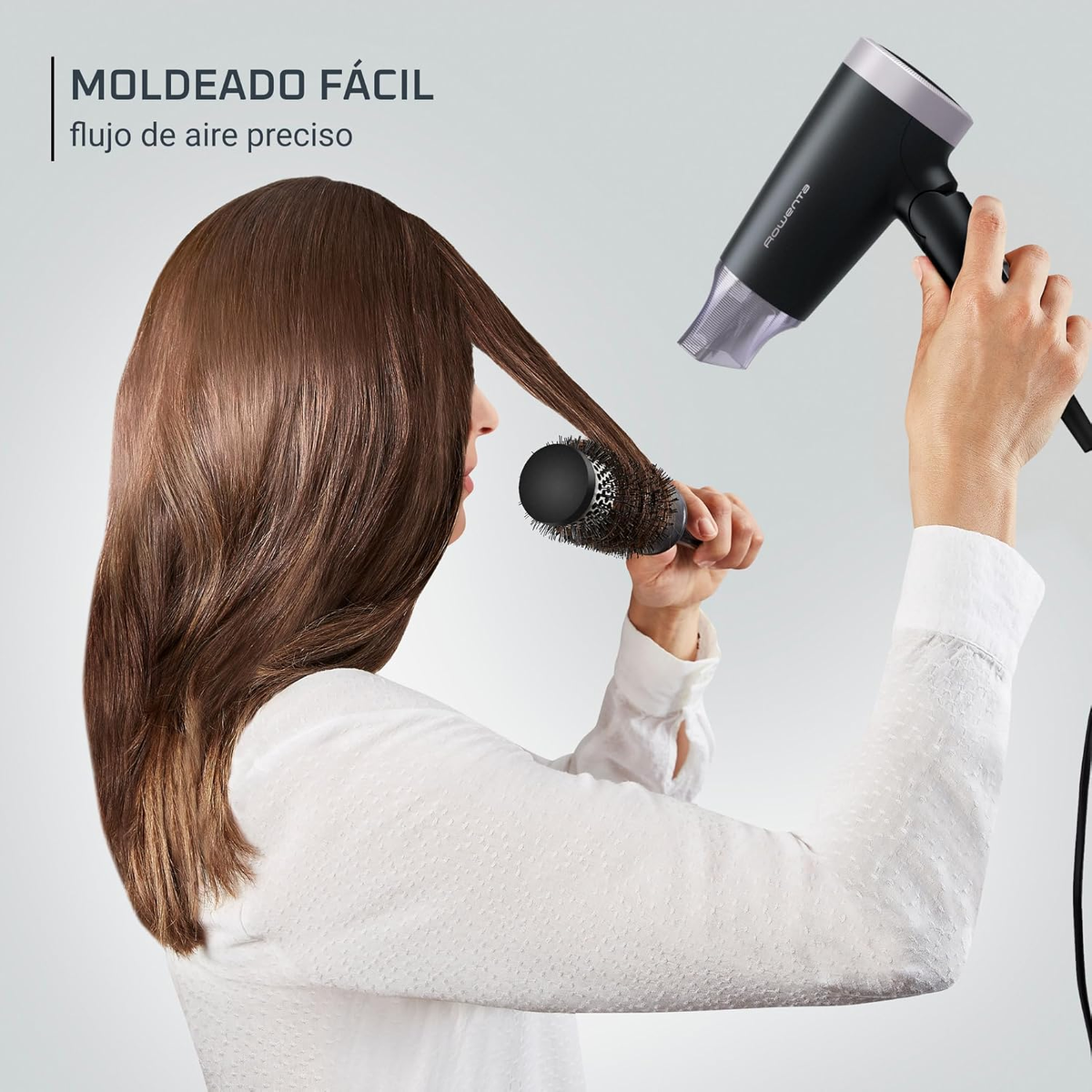 Una mujer usa un secador y un cepillo en su cabello. Secador negro, camisa blanca.