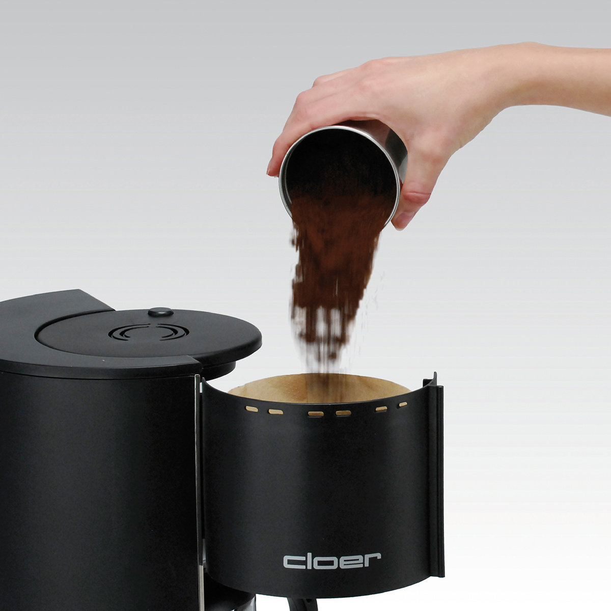 Person gießt Kaffeepulver in den Filter einer schwarzen Kaffeemaschine.