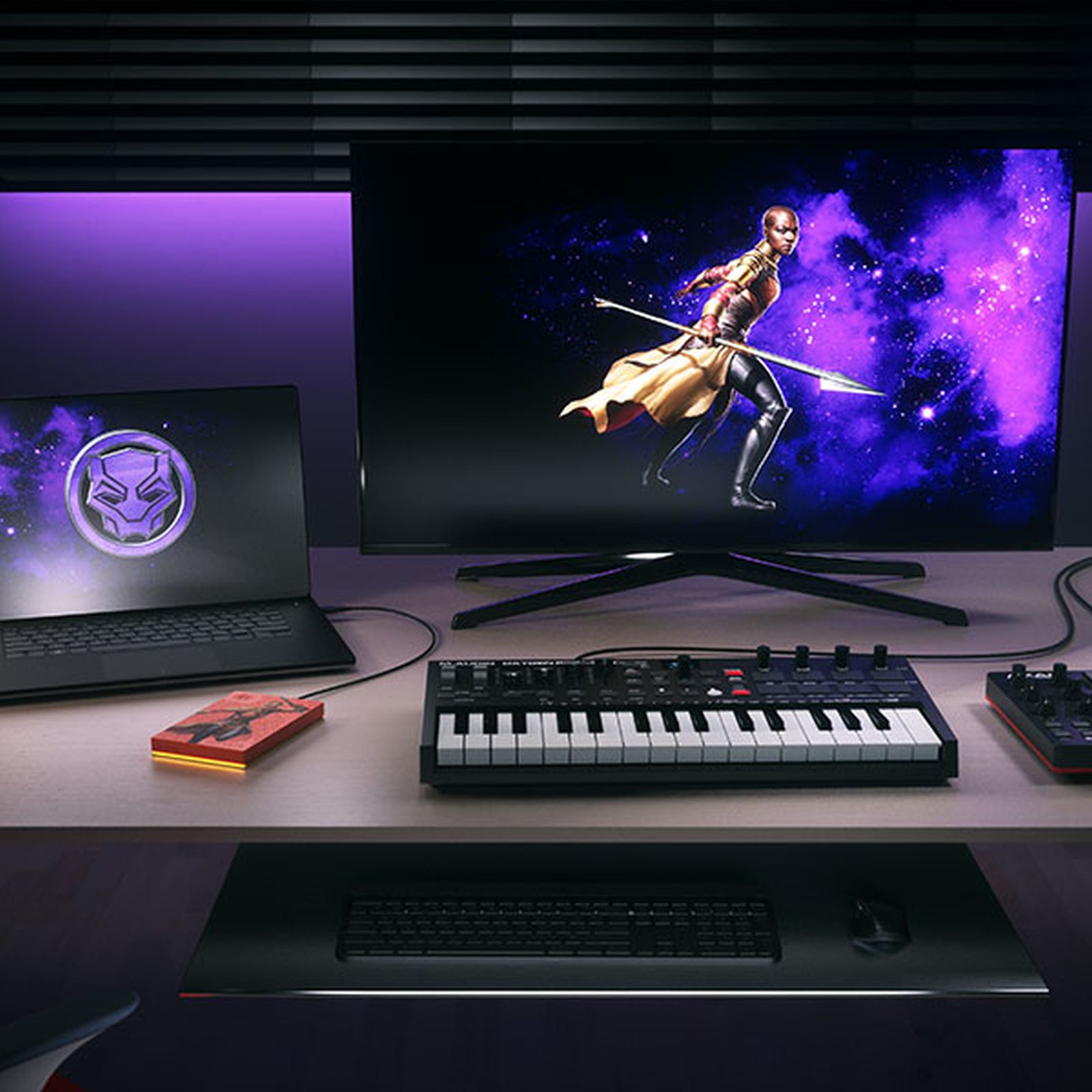 Konfiguracja biurka z monitorem wyświetlającym scenę z Czarnej Pantery, laptopem i klawiaturą.