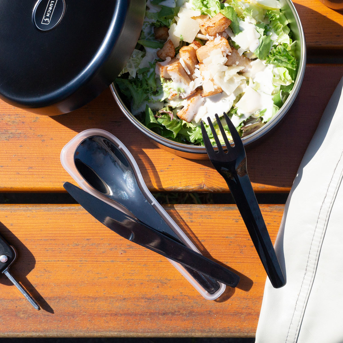 Lunchbox mit Salat, Gabel, Löffel und Messer auf einem Holztisch. Autoschlüssel dabei.