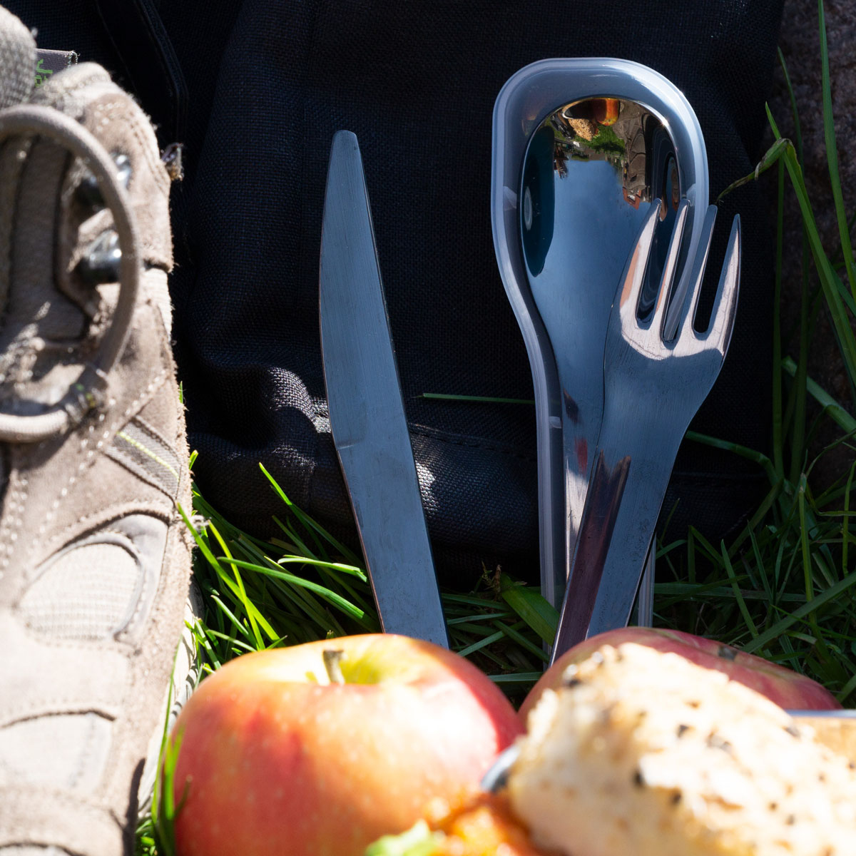 Picknick-Szene mit offenem Rucksack, Messer, Gabel, Apfel und Brot auf Gras. Wanderschuh sichtbar.