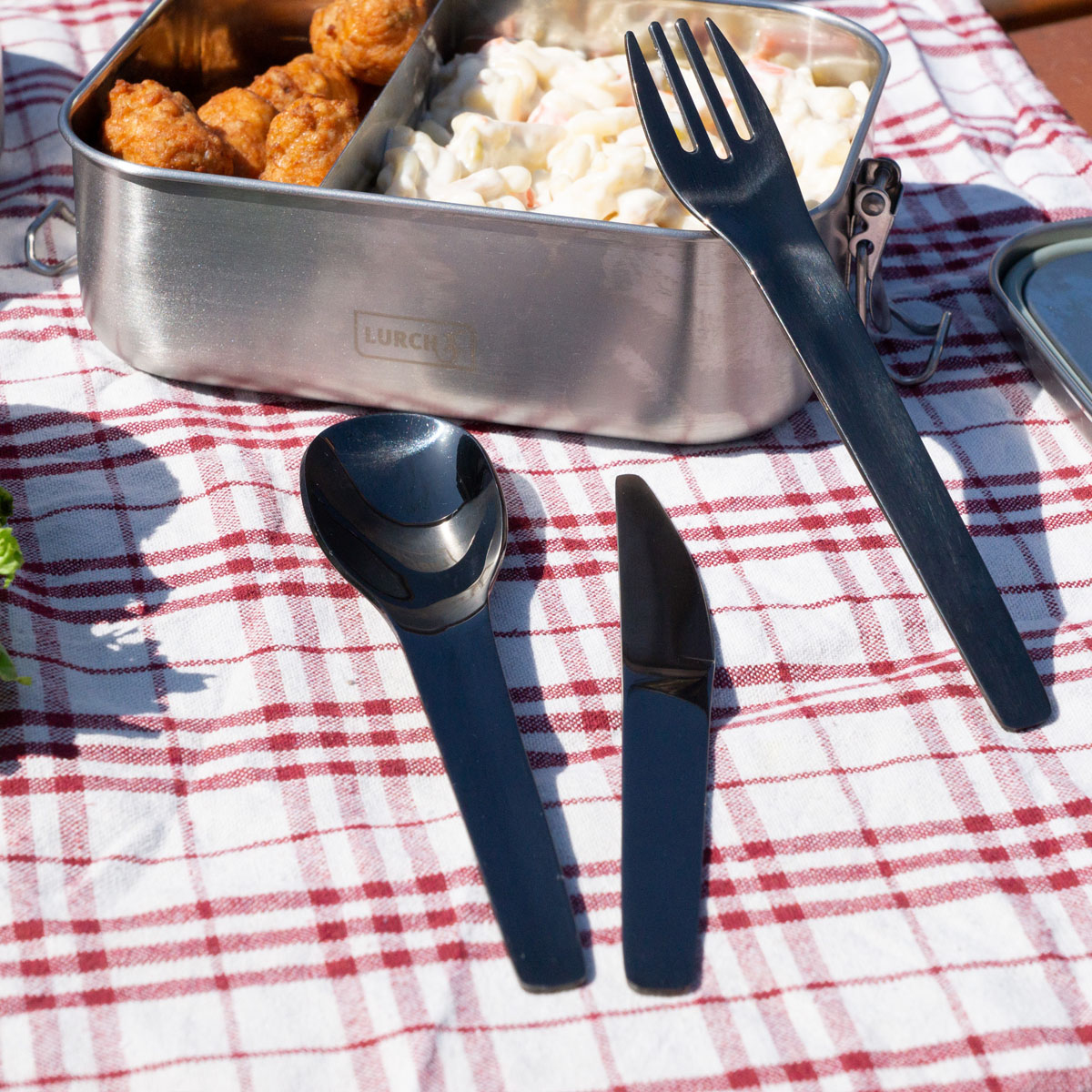 Eine Metall-Lunchbox mit Essen und Besteck. Eine rot-weiß karierte Tischdecke ist sichtbar.