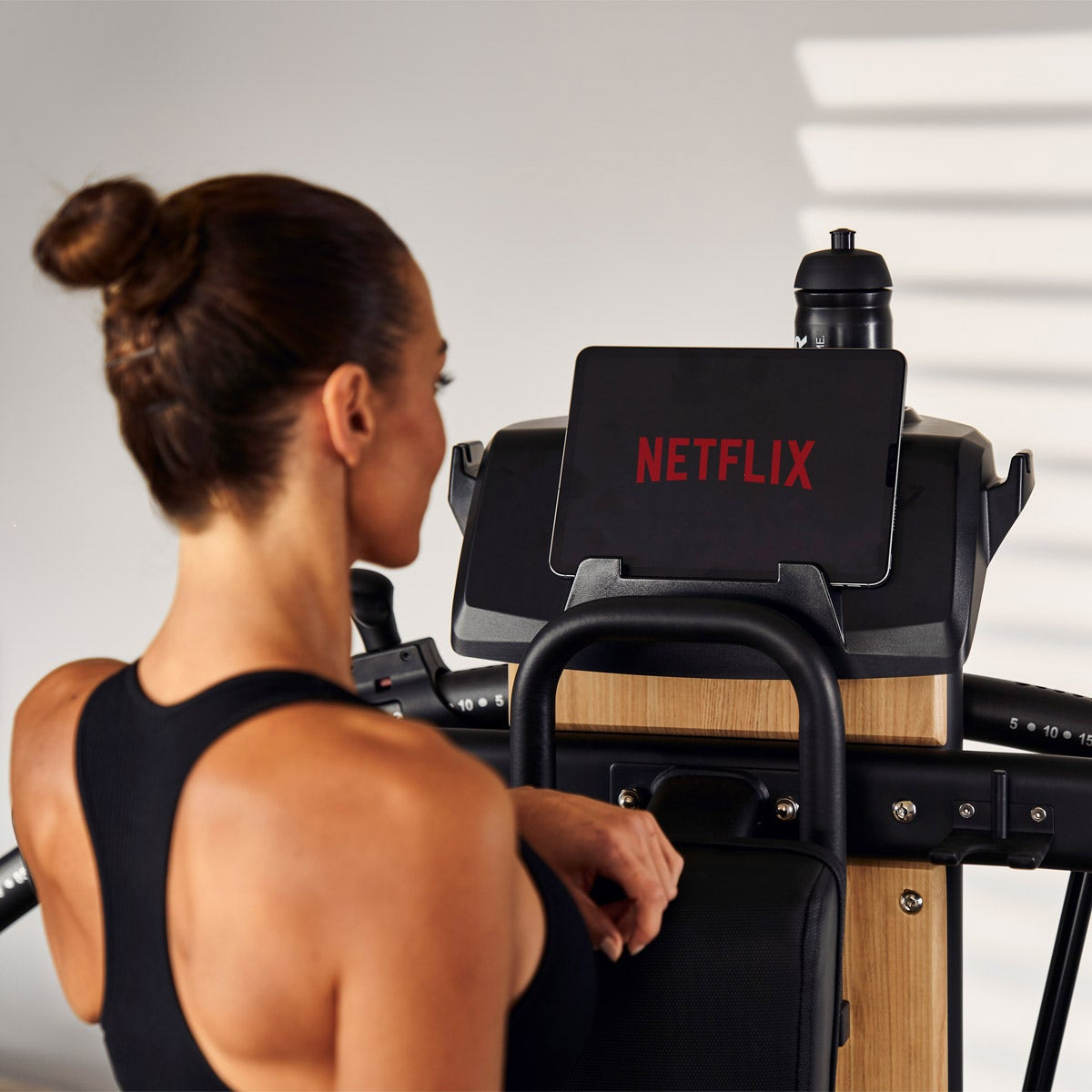 Frau benutzt ein Rudergerät mit einem Tablet, das Netflix anzeigt. Eine Wasserflasche ist in der Nähe.