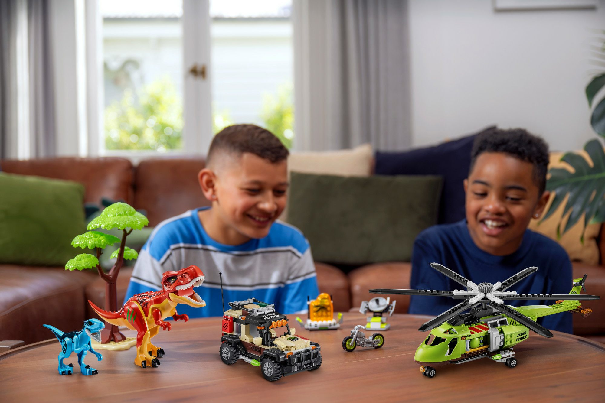 Dwóch chłopców bawiących się zabawkami Lego z dinozaurami i pojazdami na drewnianym stole, uśmiechających się do siebie.