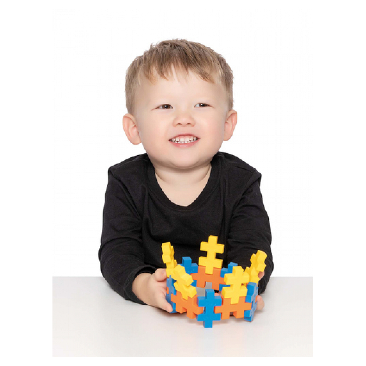 Młody chłopiec uśmiechnięty, trzyma kolorowe klocki, siedzi przy stole, białe tło.