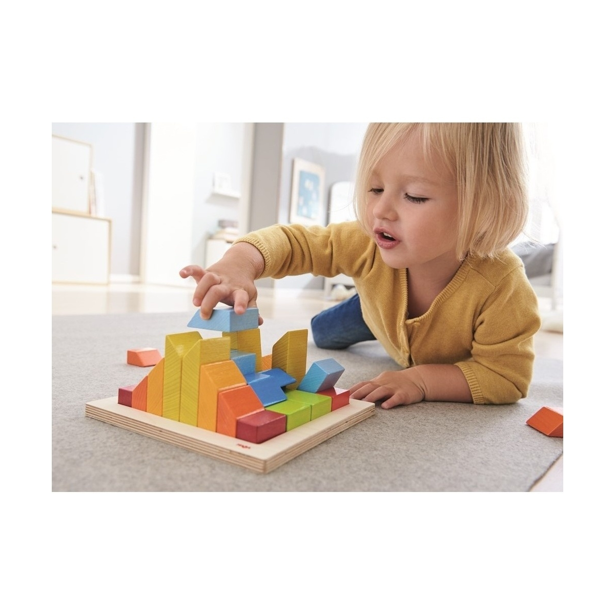 Dziecko bawi się kolorowymi drewnianymi klockami na drewnianej podstawie, na szarym dywanie.