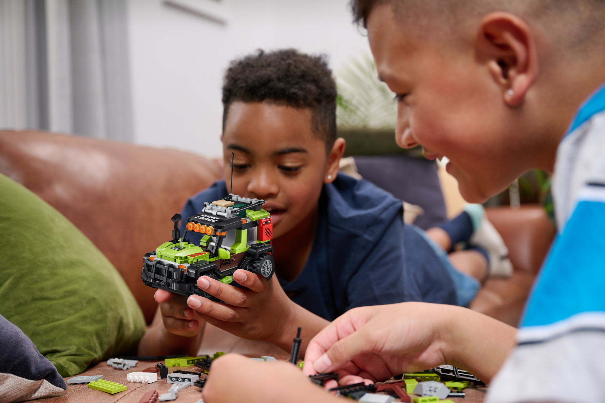 Due ragazzi che costruiscono un veicolo Lego. Uno è di carnagione scura e l'altro è di carnagione chiara. Sono su un divano marrone.