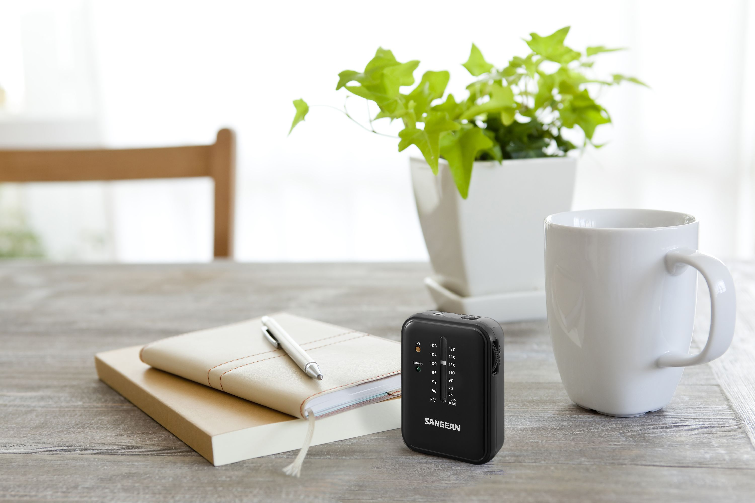 Czarne radio na drewnianym stole, obok notatnika, długopisu, rośliny i białego kubka.