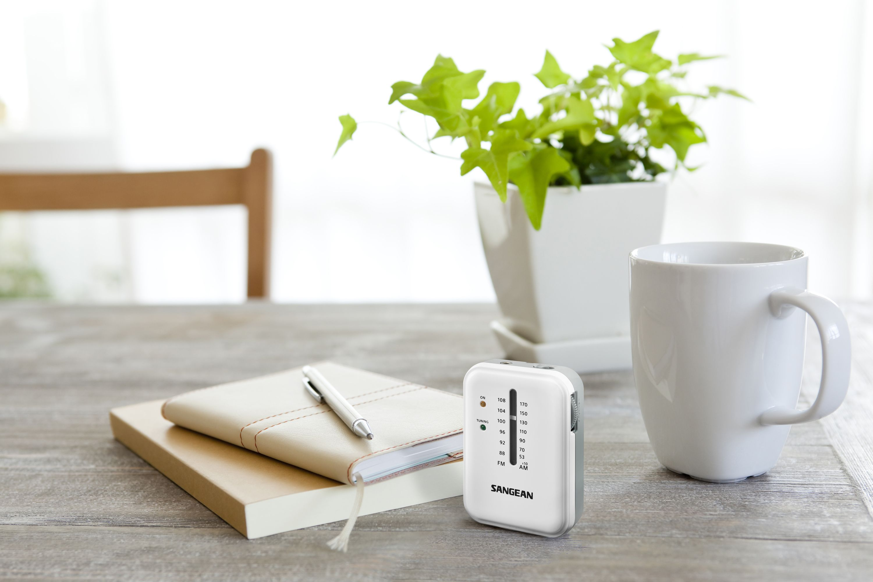 Białe radio na drewnianym stole z rośliną, notatnikiem, długopisem i kubkiem. Jasne, neutralne oświetlenie.