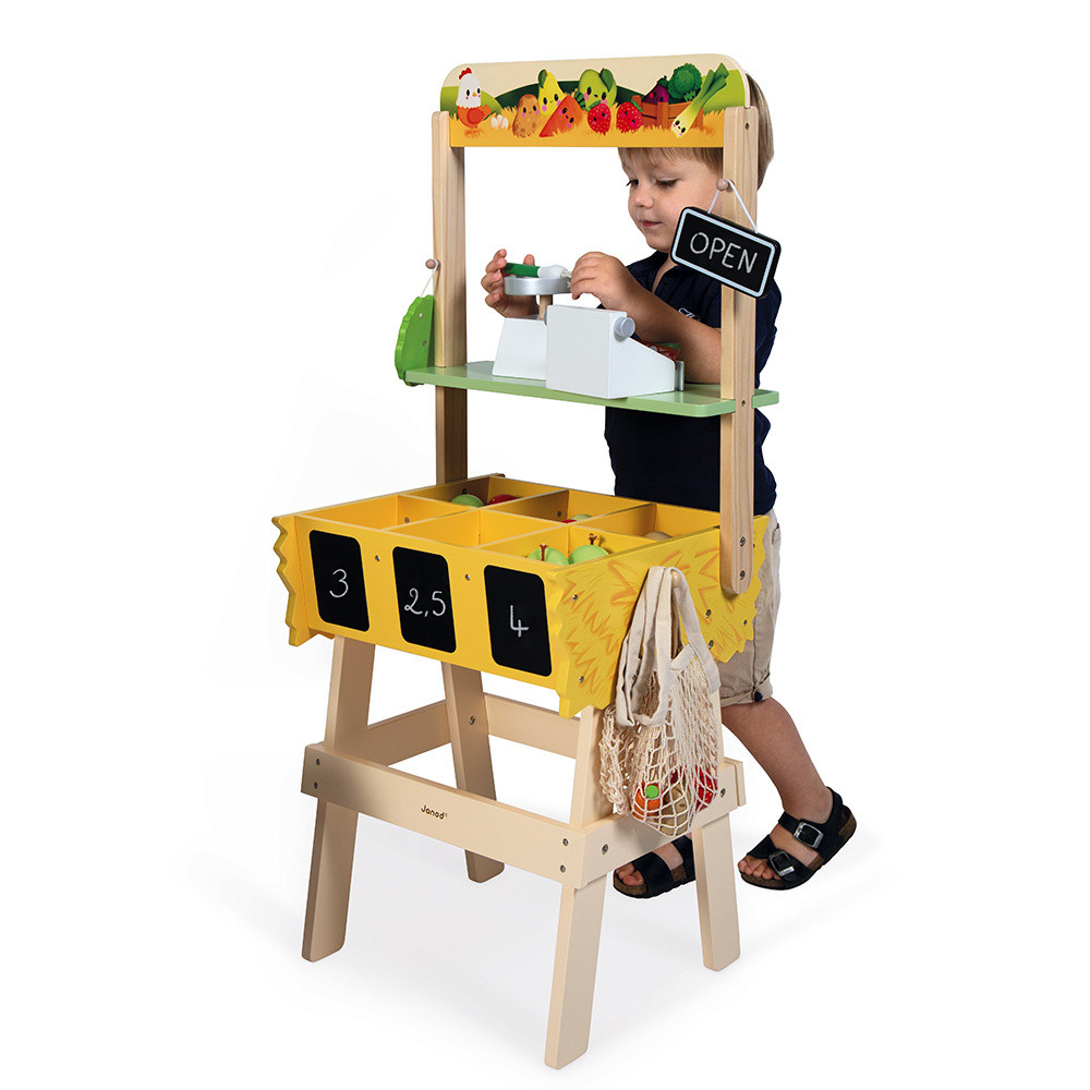 Ein Kind spielt mit einem Holzspielzeug-Obststand. Es hat Früchte und ein Schild mit der Aufschrift OPEN.