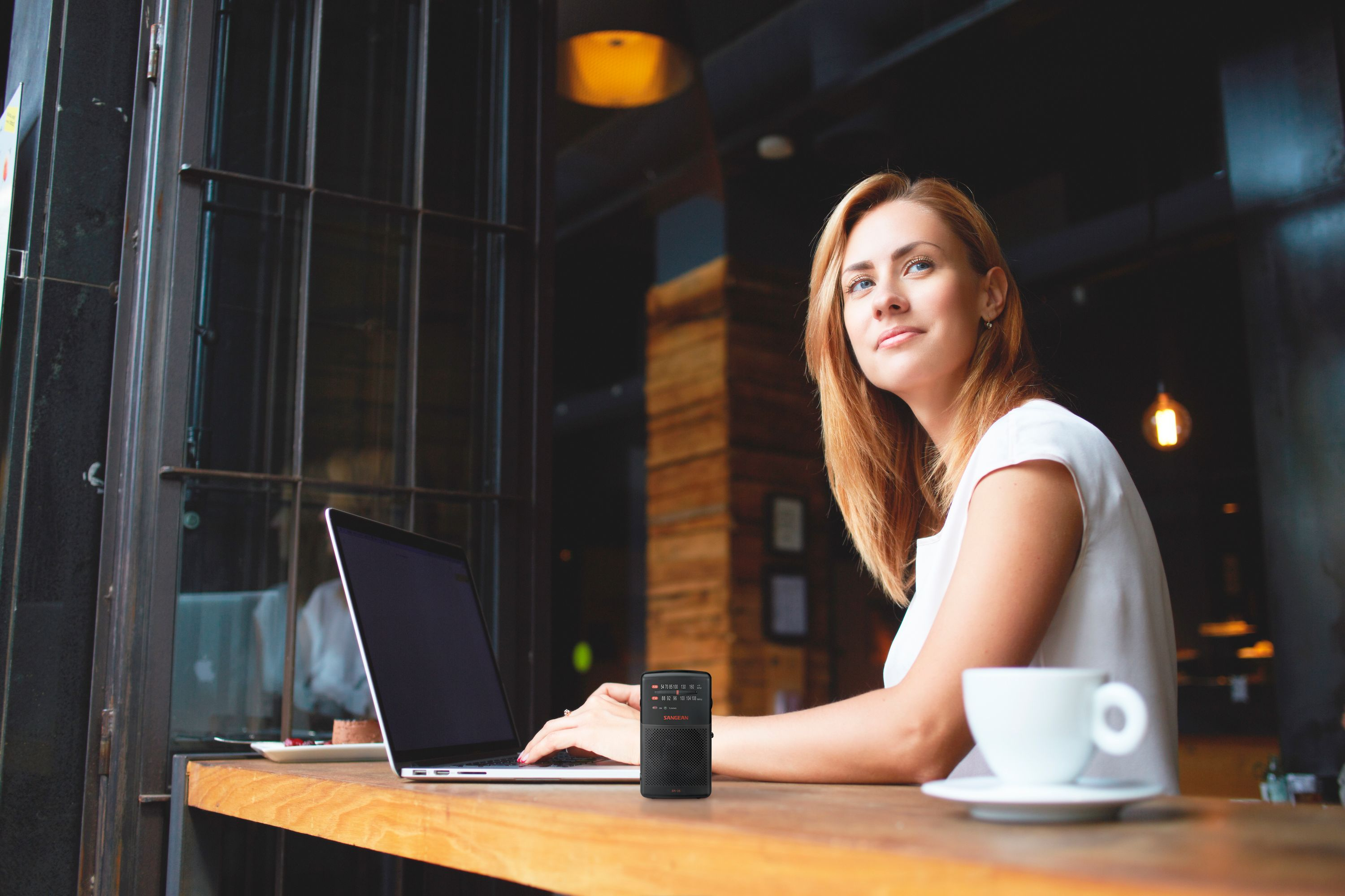 Woman at a table with a laptop, radio, and coffee cup, near a window.