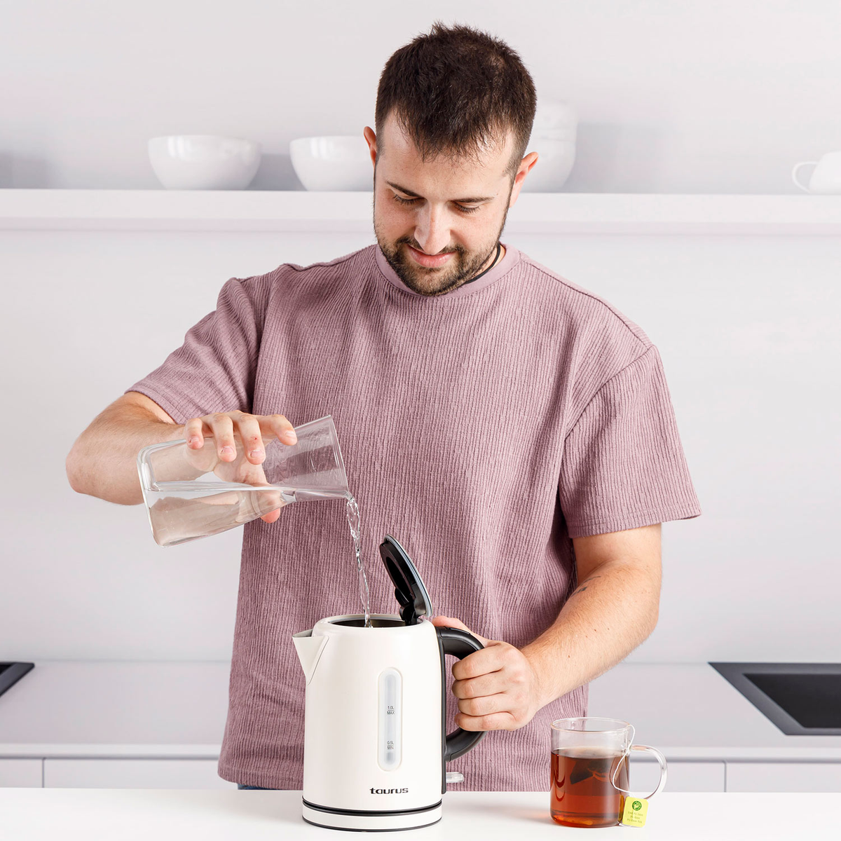 Un uomo versa acqua in un bollitore bianco, con una tazza di tè visibile.