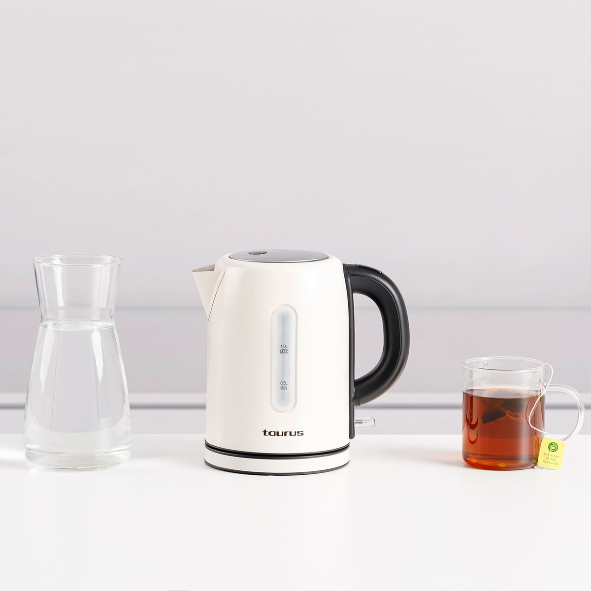 Bollitore elettrico bianco, caraffa d'acqua in vetro e un bicchiere di tè su una superficie bianca.