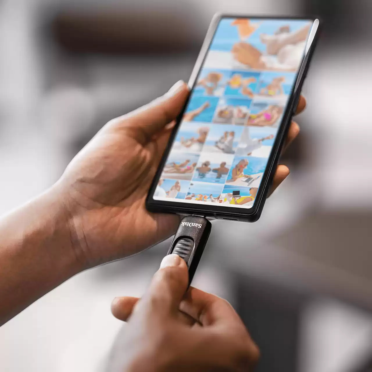 Dłoń trzyma czarny pendrive SanDisk USB-C, podłączony do telefonu wyświetlającego zdjęcia z wakacji.