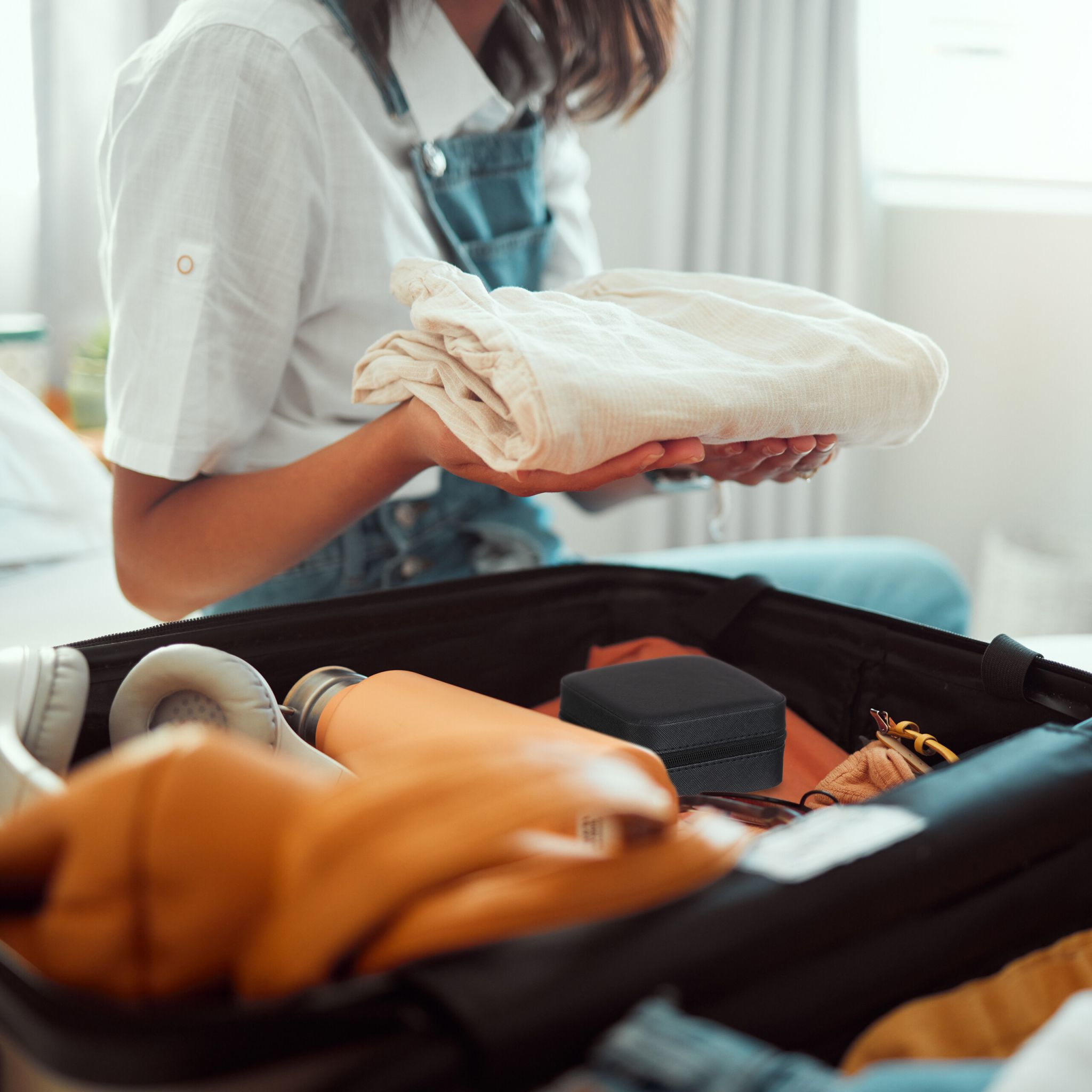 Person packt Koffer mit Kleidung. Eine schwarze Box und andere Gegenstände sind sichtbar.