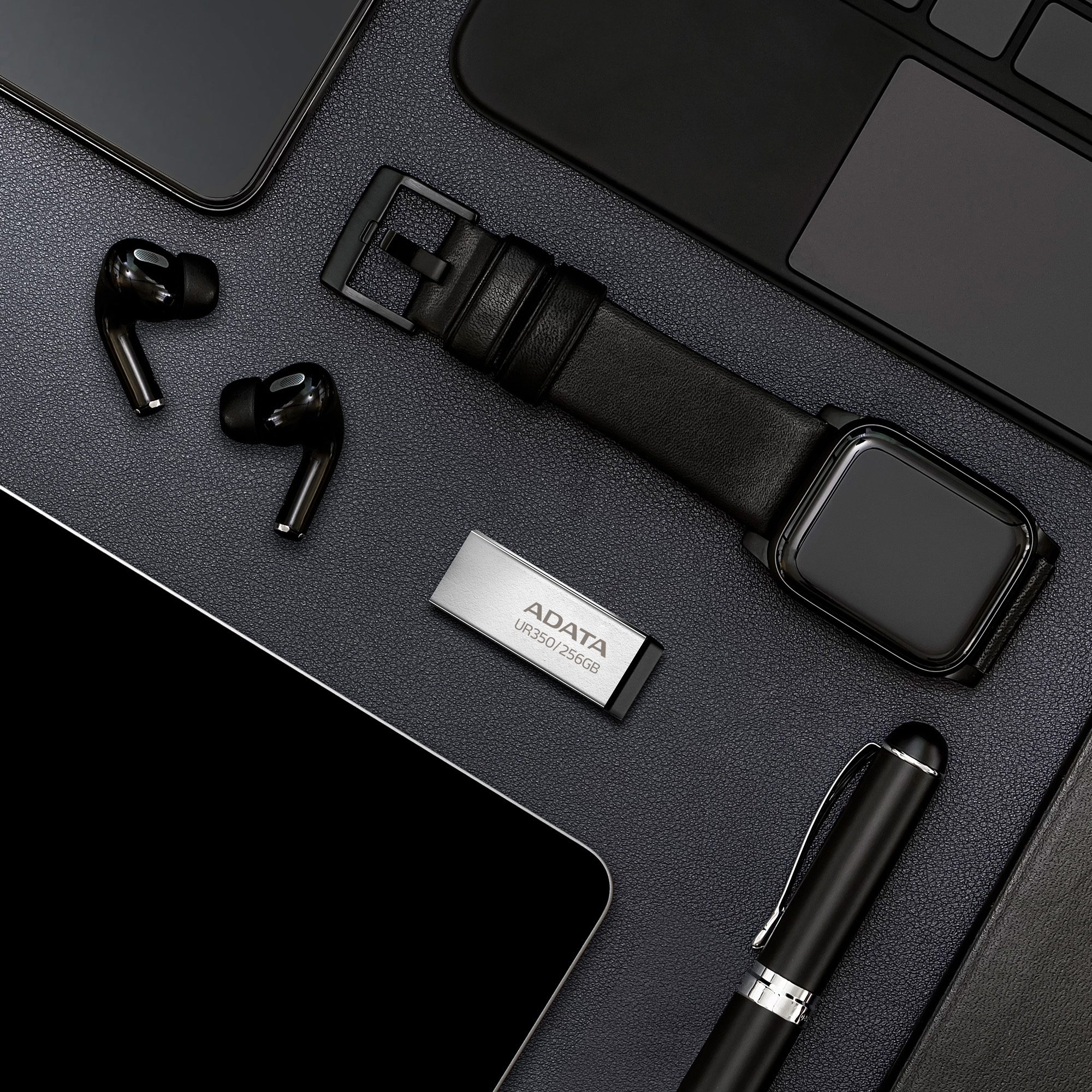 Czarne miejsce pracy z gadżetami technologicznymi: słuchawki, smartwatch, pendrive, długopis, tablet i laptop.