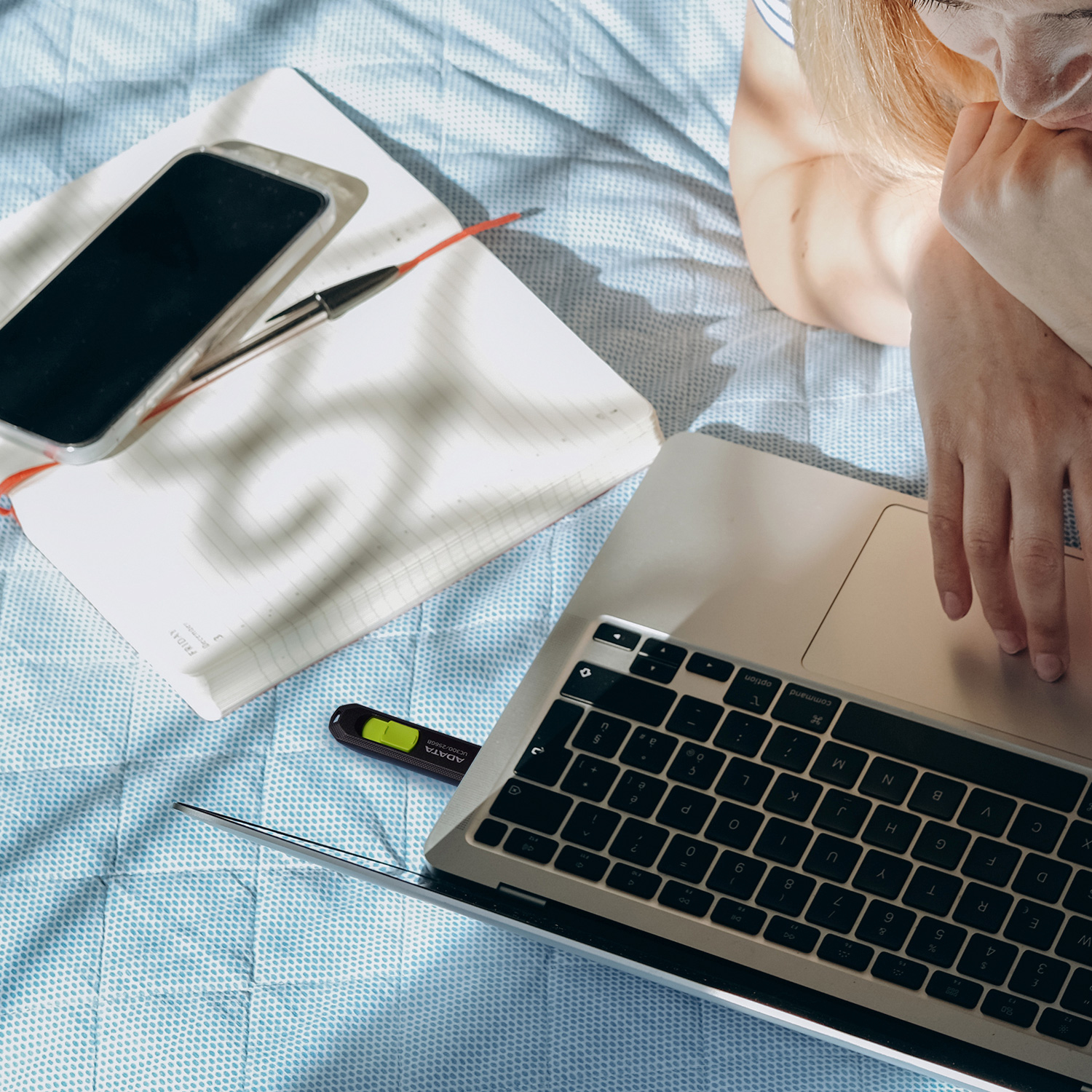 Laptop, smartfon, długopis i notatnik na niebieskim łóżku w wzory. Widoczne ramię osoby.
