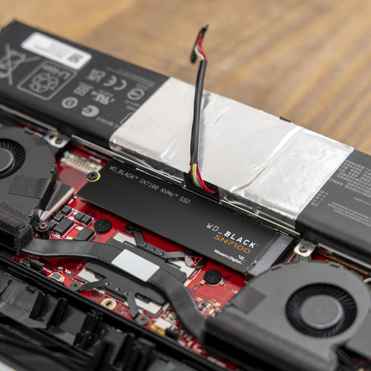 Wnętrze laptopa z dyskiem SSD, czerwoną płytką drukowaną, wentylatorem, baterią i przewodami. Dysk SSD to WD BLACK SN7100.