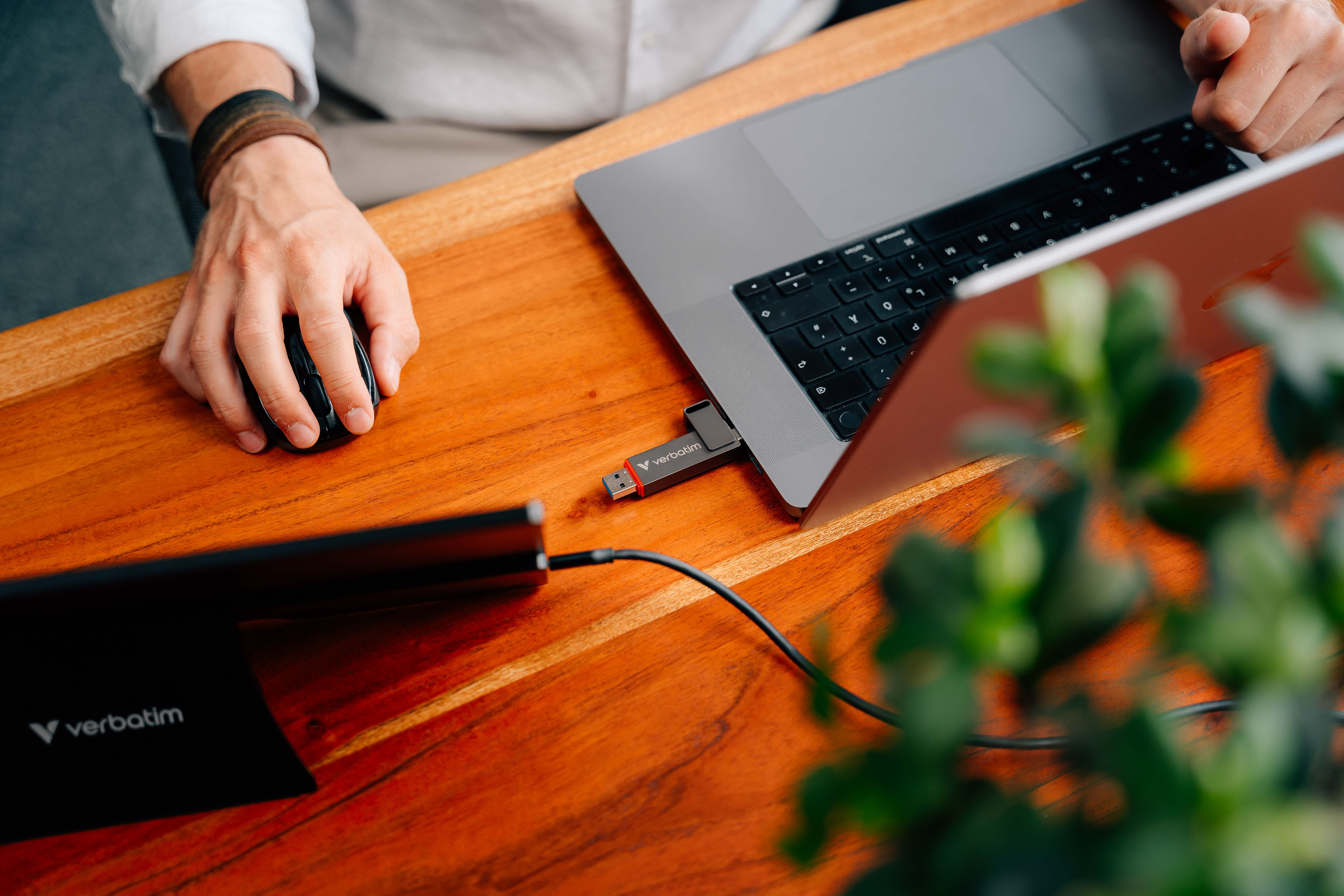 Osoba korzystająca z laptopa i myszy na drewnianym stole. Pamięć USB jest podłączona do laptopa. Jest roślina.