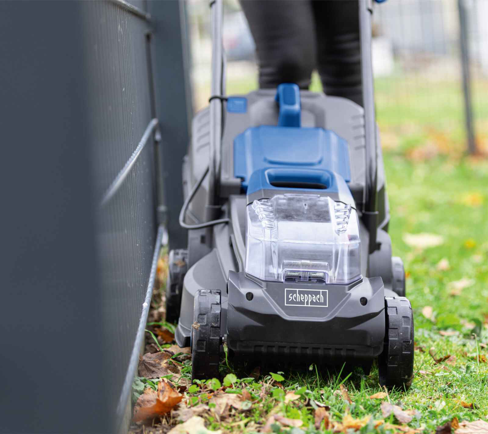 Een blauw-grijze grasmaaier die gras maait naast een hek.