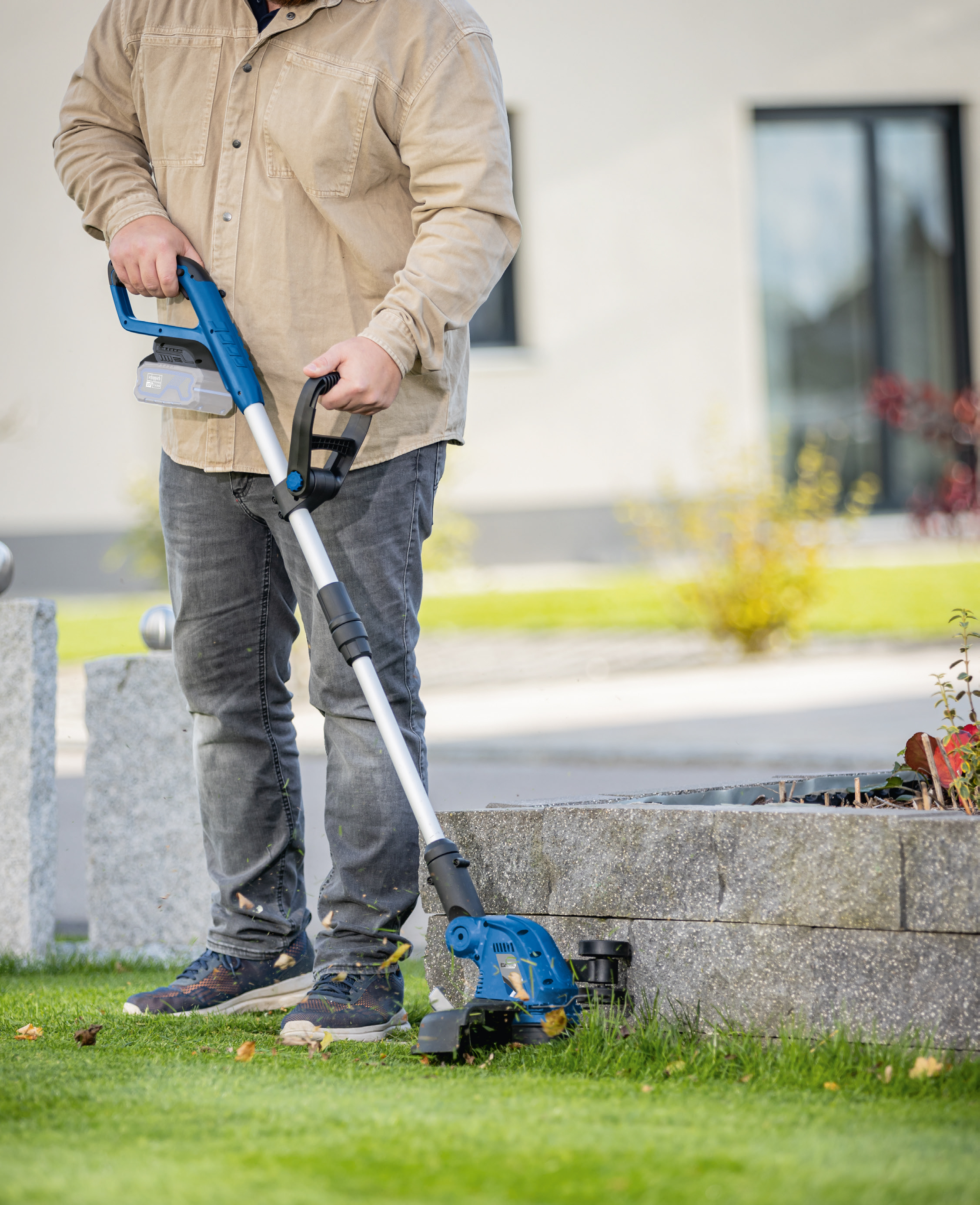 Eine Person benutzt einen blau-schwarzen Trimmer in der Nähe einer Steinmauer auf grünem Gras.