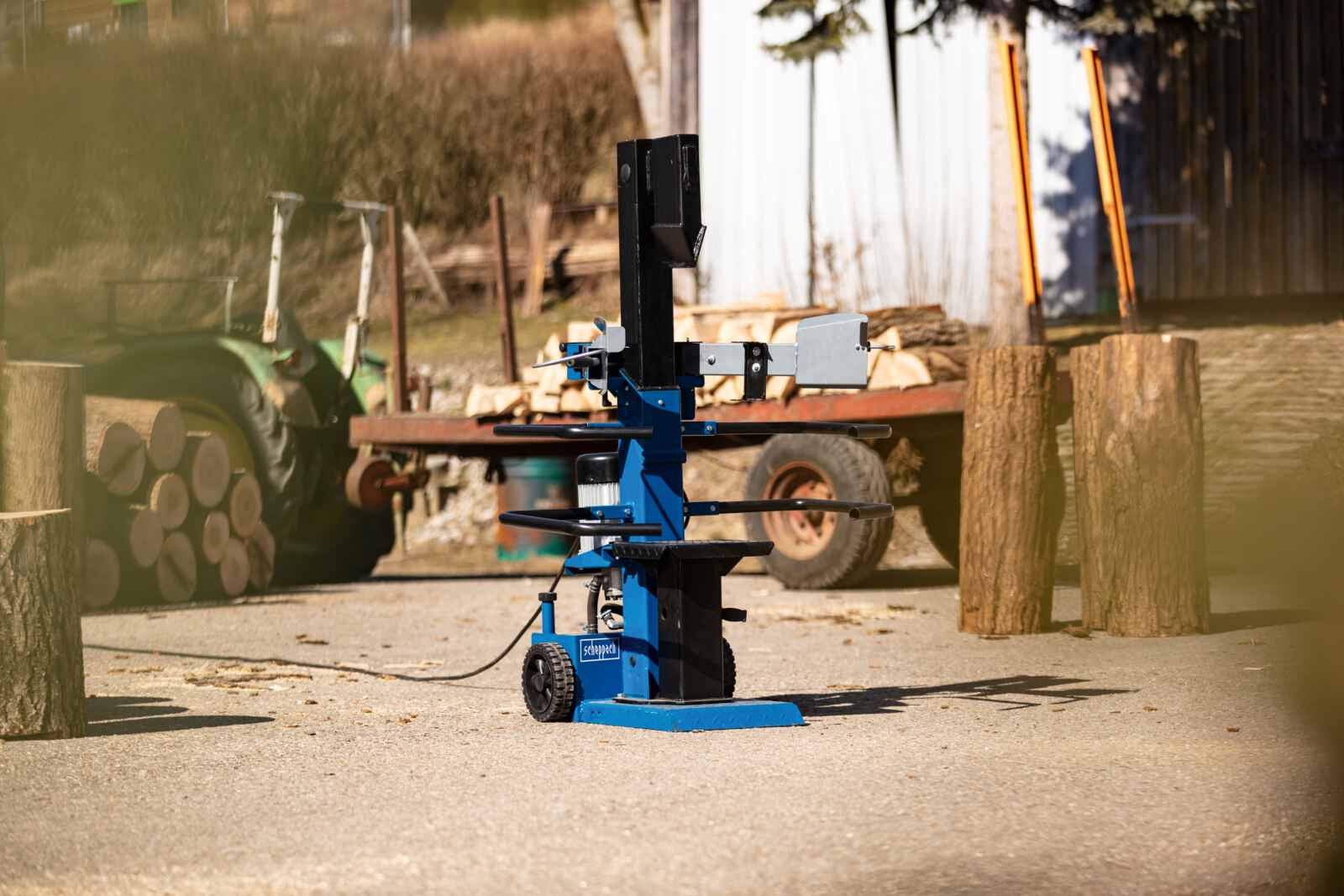 Een blauwe houtklovermachine op een betonnen ondergrond, met houtblokken op de achtergrond.