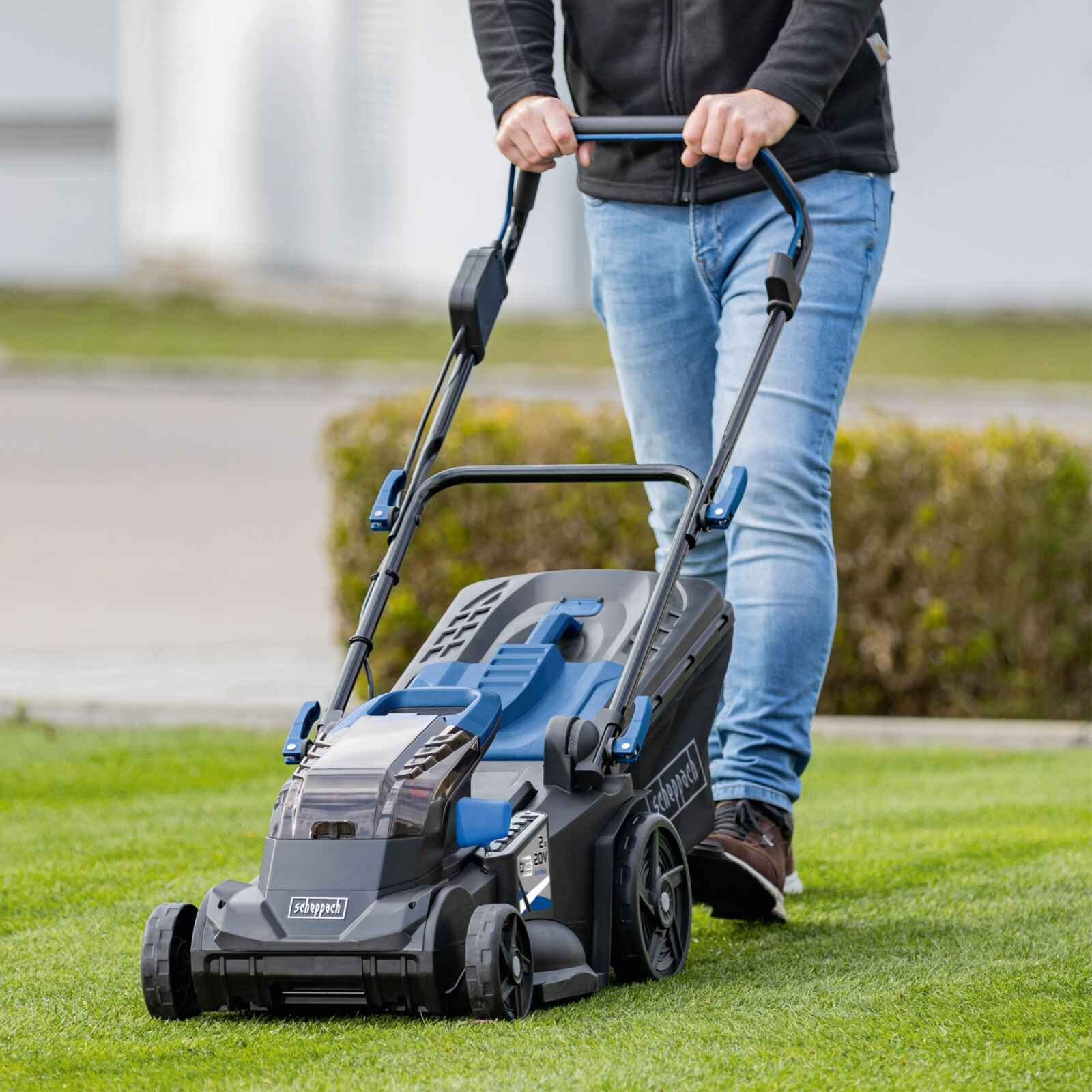 Eine Person mäht mit einem blau-schwarzen Rasenmäher auf einer Rasenfläche in sonniger Umgebung den Rasen.