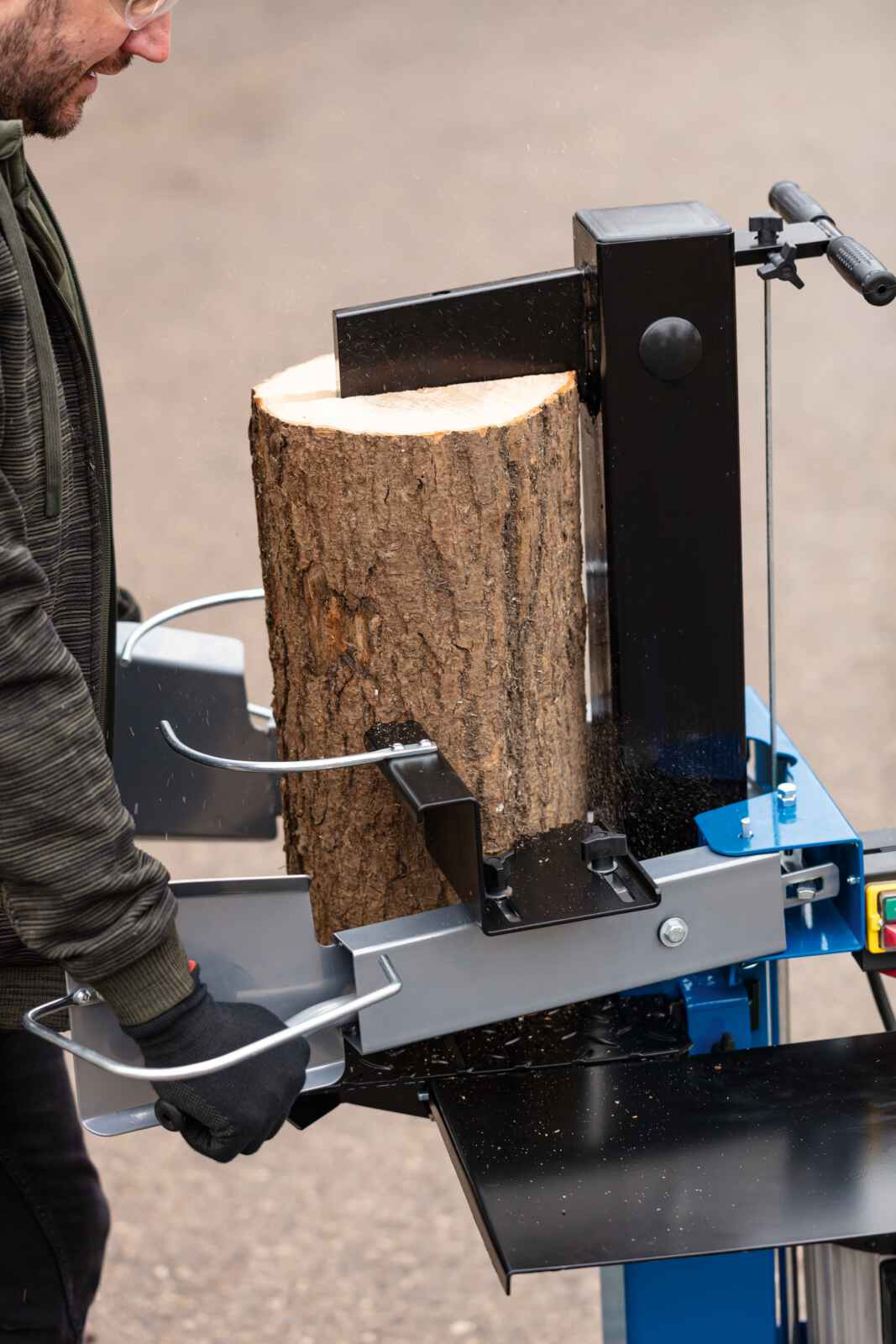 Een persoon gebruikt een houtklover. Een blok hout wordt gespleten. De machine is blauw en zwart.