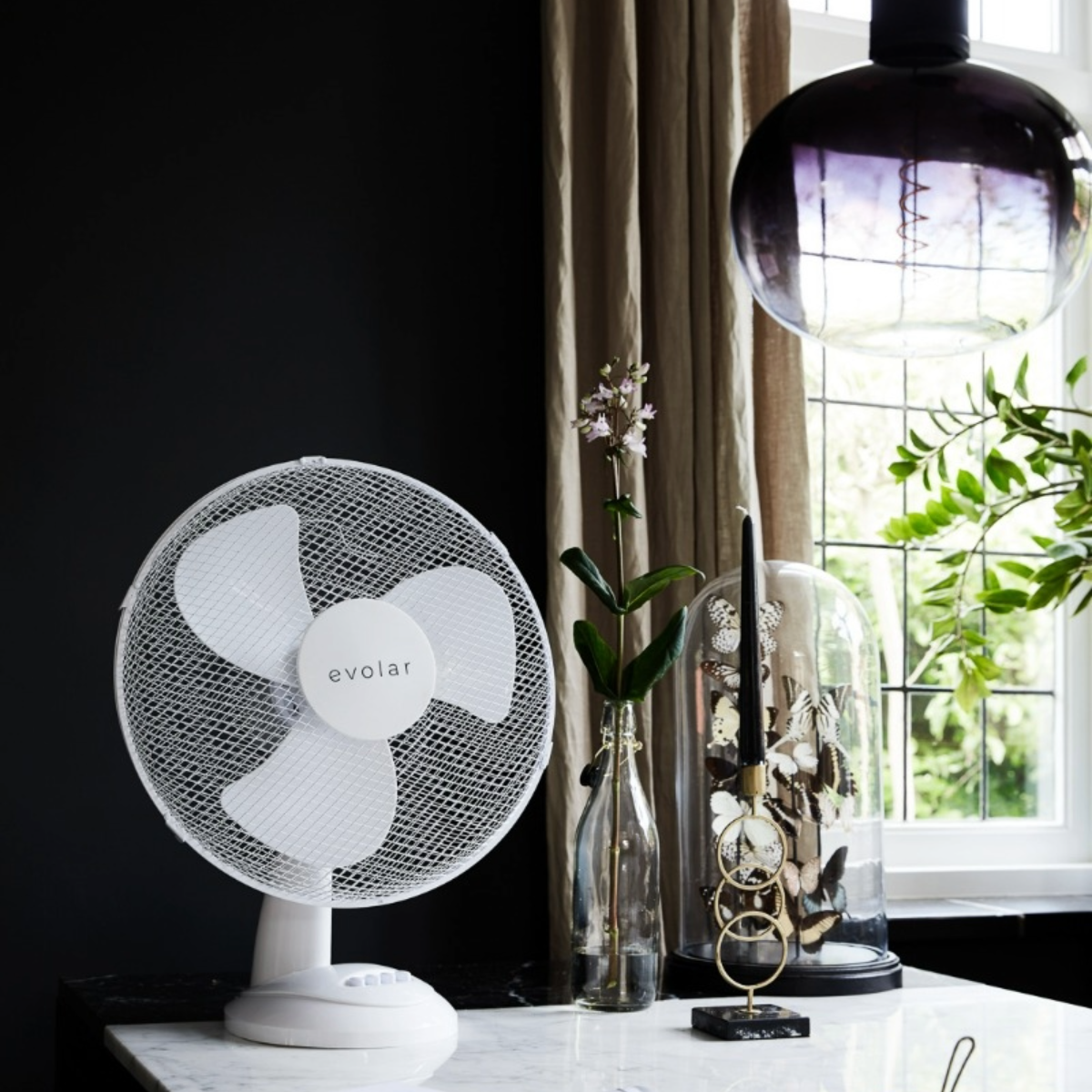 A white desk fan with three blades, near a window with a view. Decorative items are on the desk.
