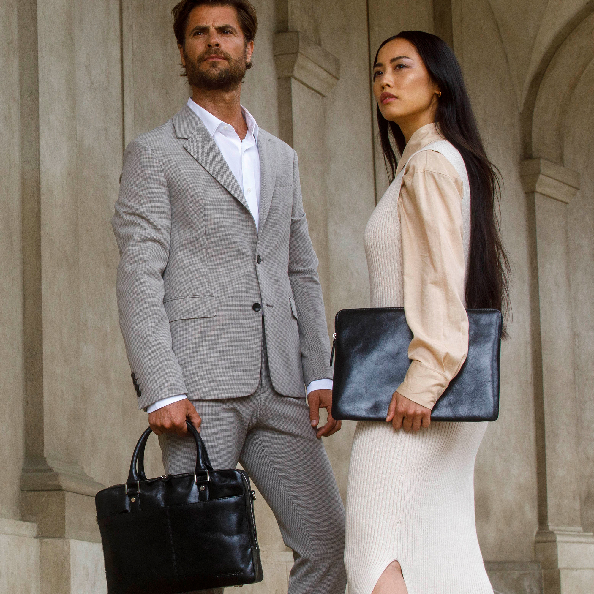 Un uomo in giacca e cravatta e una donna in abito sono fuori. Entrambi tengono borse nere. Edifici sullo sfondo.