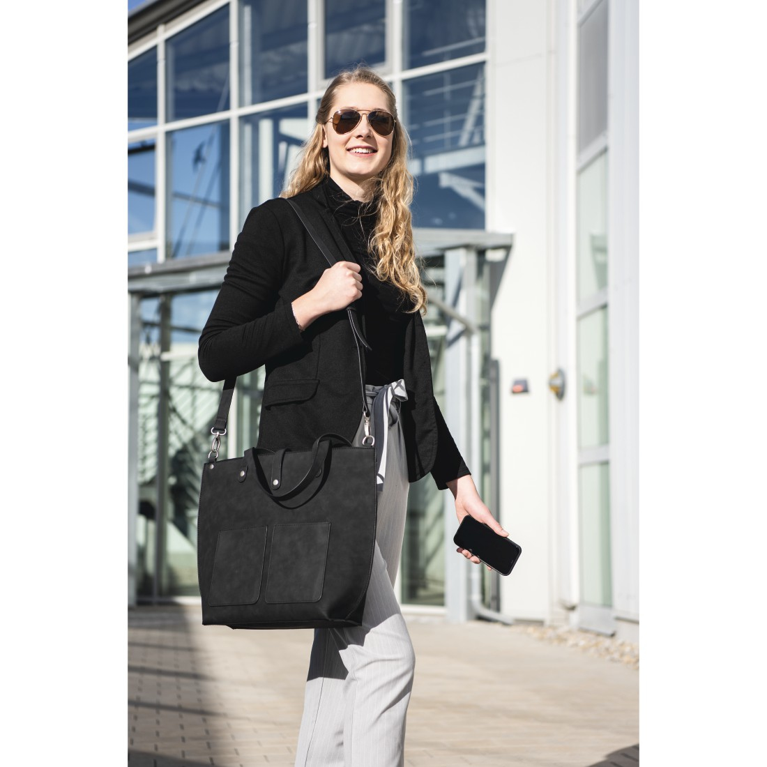 Donna con occhiali da sole, blazer e telefono, cammina vicino a un edificio, con borsa.