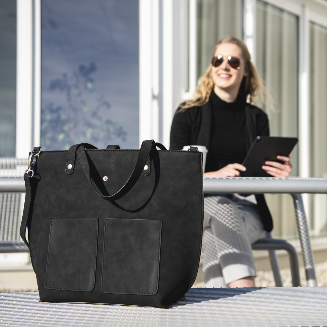 Donna con occhiali da sole seduta, tiene un tablet, borsa nera su un tavolo.