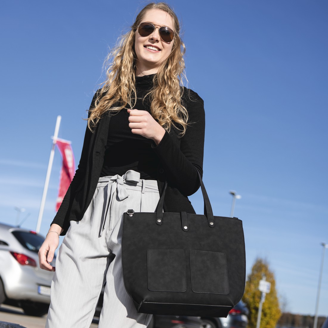 Donna con occhiali da sole e borsa, sorridente davanti a un parcheggio e cielo blu.