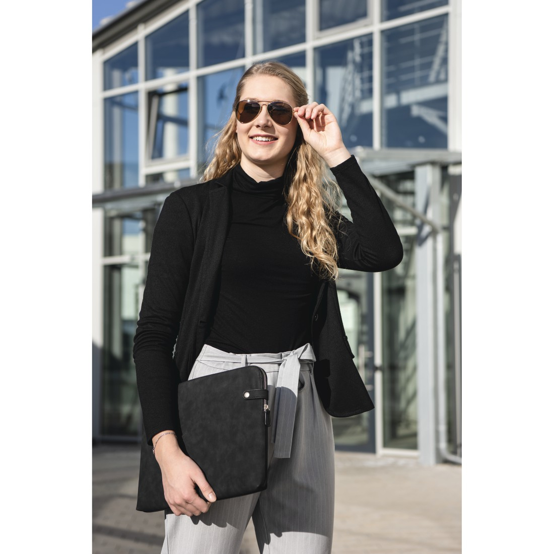 Blondynka w okularach przeciwsłonecznych i czarnej kurtce trzymająca etui na laptopa, uśmiechnięta.